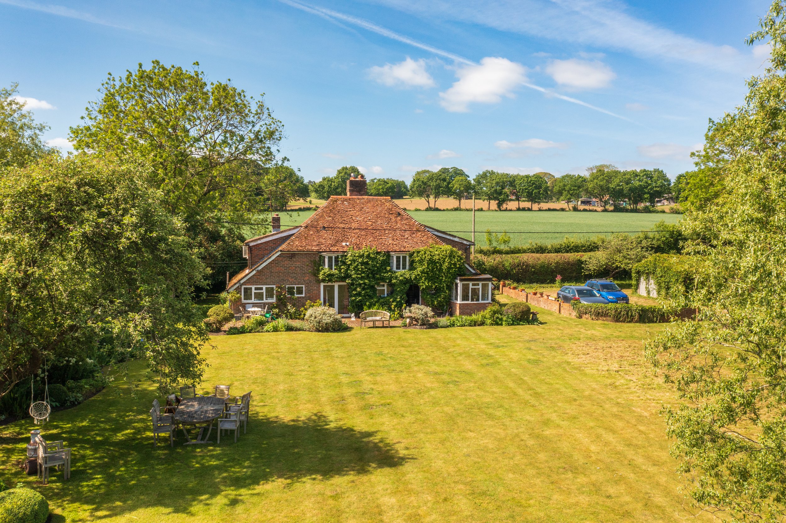 A large brick house with a red tile roof, surrounded by lush green trees and a well-maintained lawn, with a table and chairs in the yard, and parked cars nearby, under a blue sky with a few clouds.