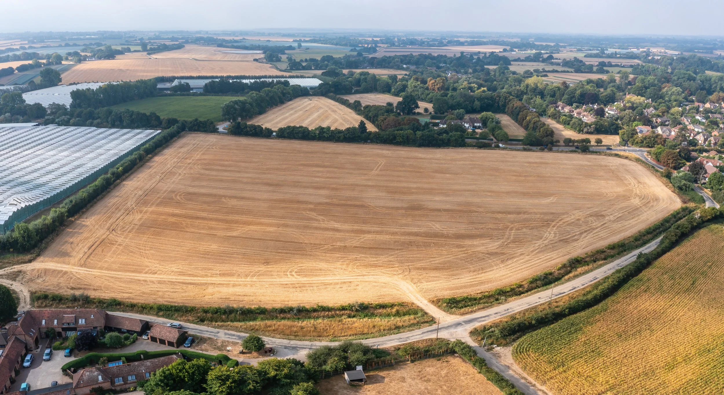 Aerial view of farmland with large, golden brown cultivated field surrounded by roads, trees, and nearby houses.