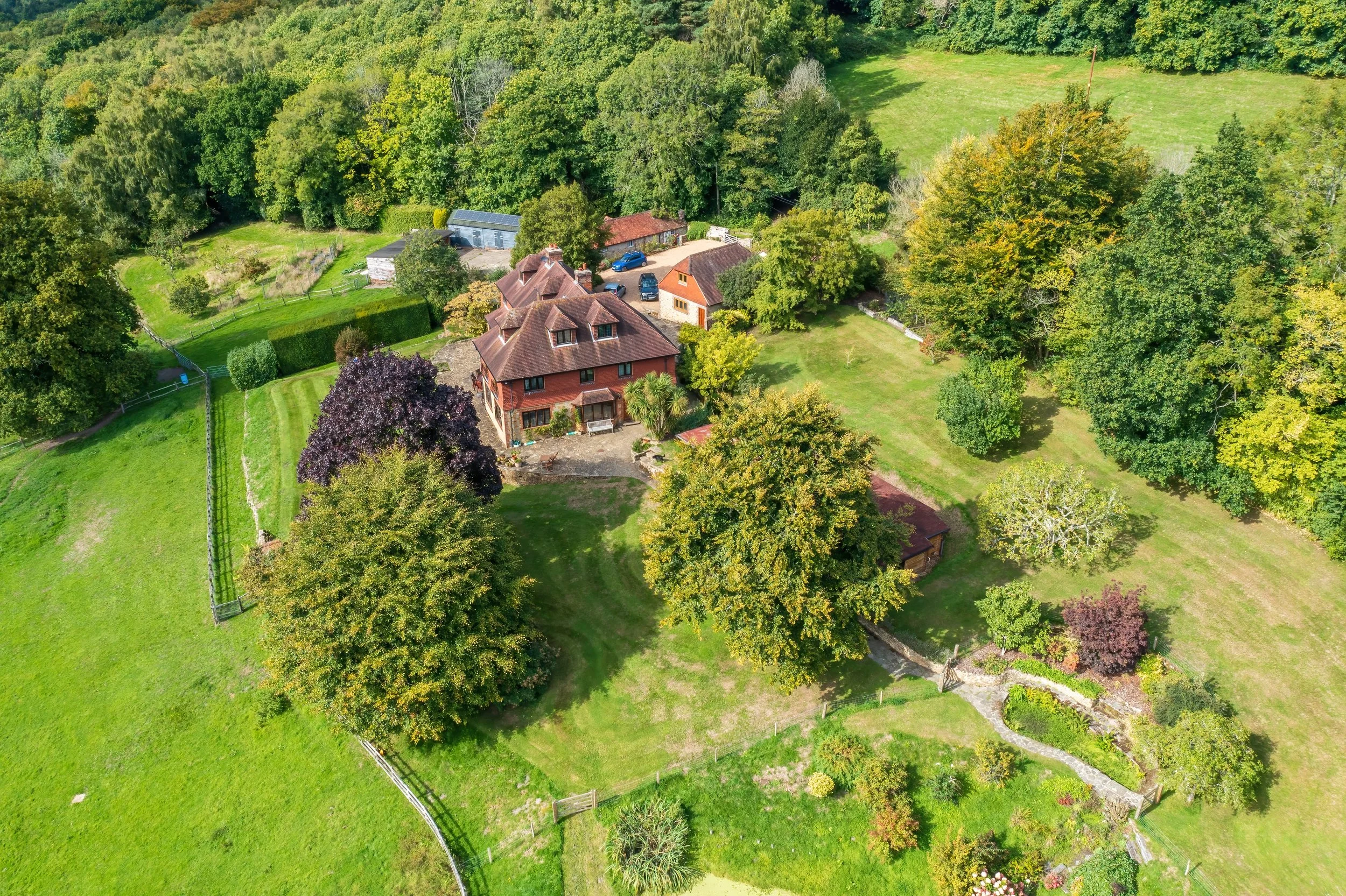 An aerial view of a large red brick house with multiple gabled roofs, surrounded by lush green trees, manicured lawns, and a garden with pathways, fences, and various shrubs.