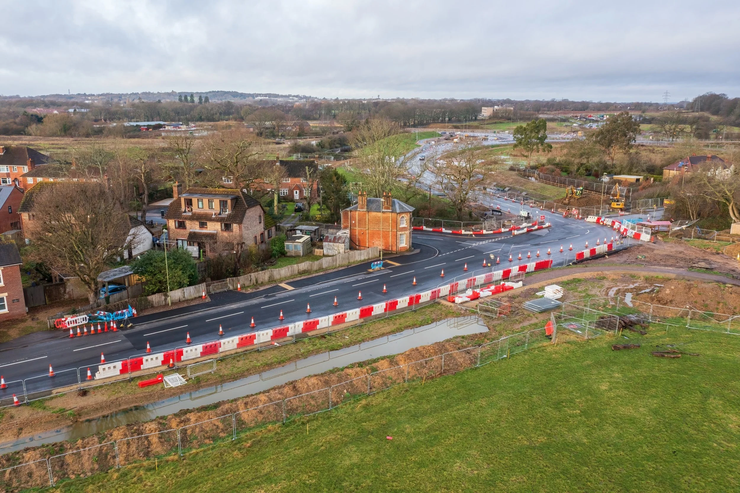 An aerial view of a road construction site with orange and white barriers, traffic cones, and construction equipment, with residential houses and green fields in the background.