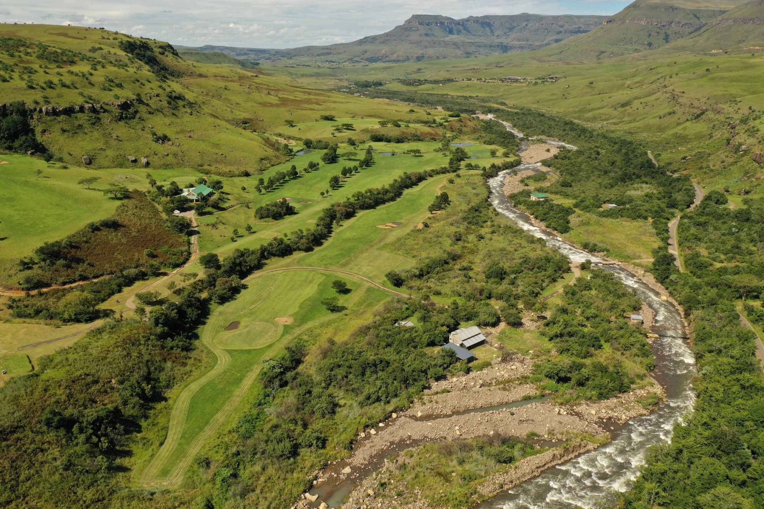 Aerial view of a lush green landscape with a winding river on the right and a golf course on the left, surrounded by hills and mountains in the background.