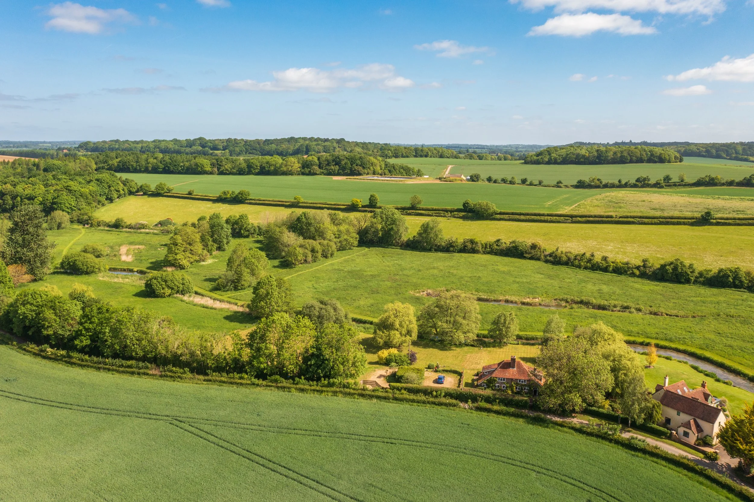 Aerial view of green fields, trees, and houses in the countryside under a partly cloudy sky.