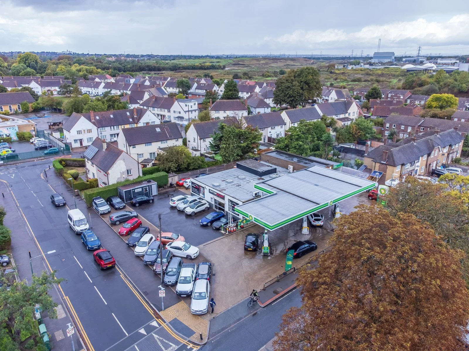 Aerial view of a petrol station with parked cars, surrounded by a residential neighborhood with houses and trees in the background.