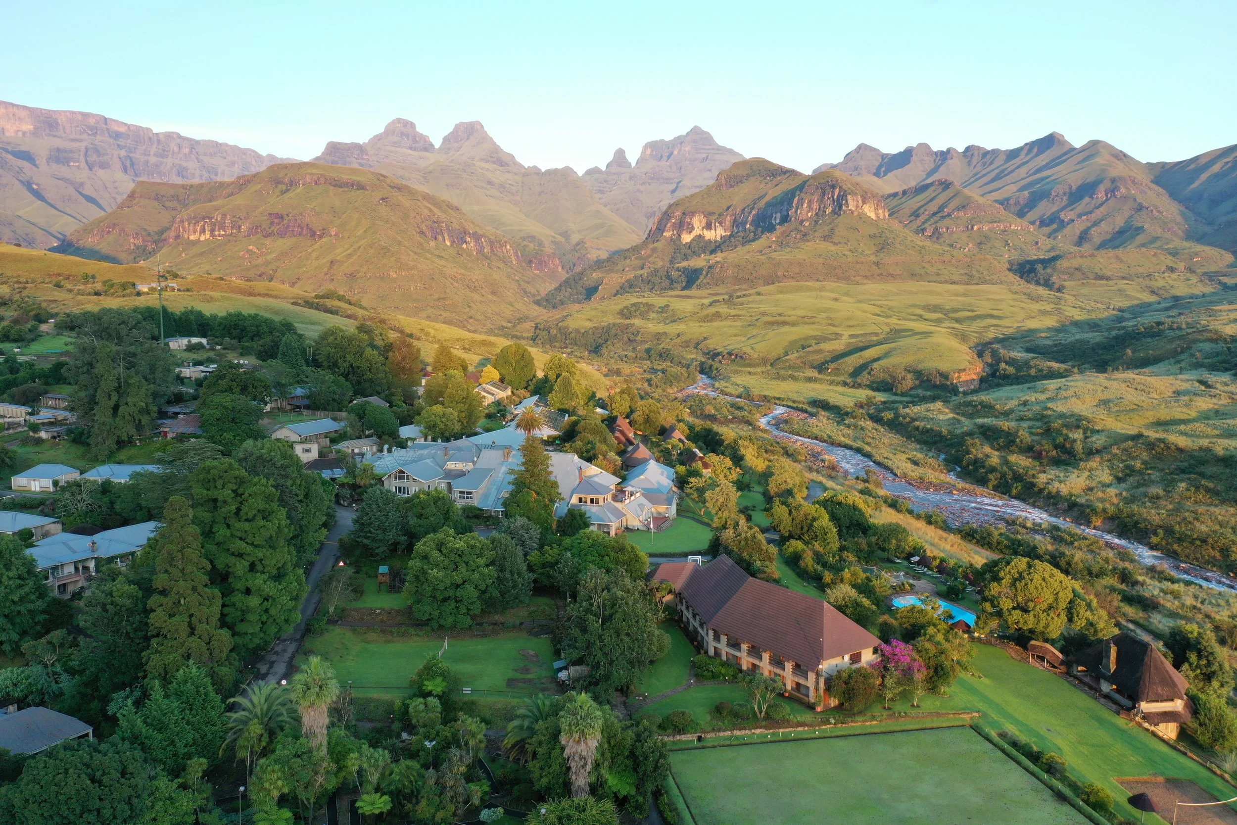 Aerial view of a lush green village with houses, trees, and a swimming pool, surrounded by mountains and a river.