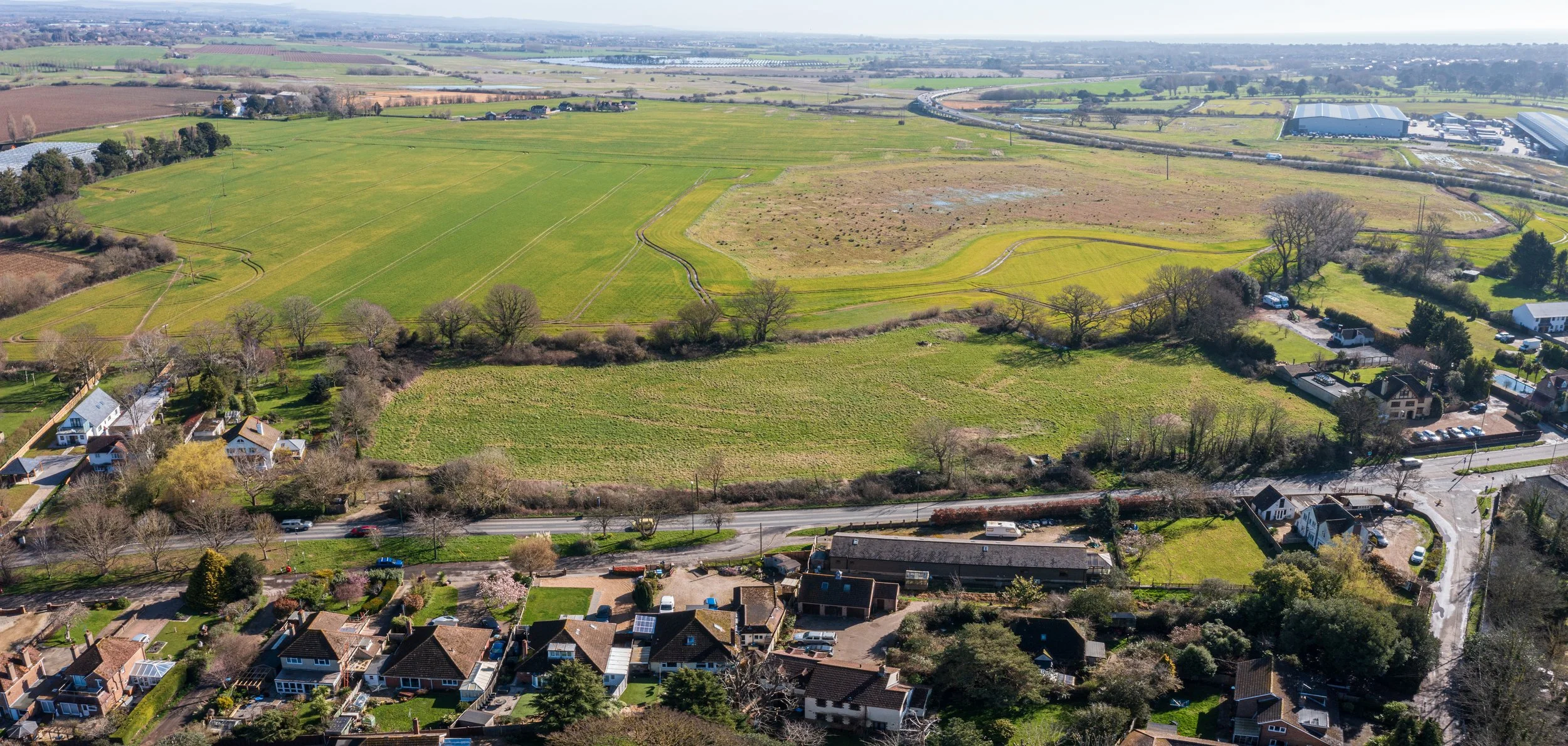 Aerial view of a suburban neighborhood with houses, trees, and green yards in the foreground. Beyond the neighborhood, there are open fields and farmland, with winding dirt paths and a few small water patches. In the background, industrial buildings 