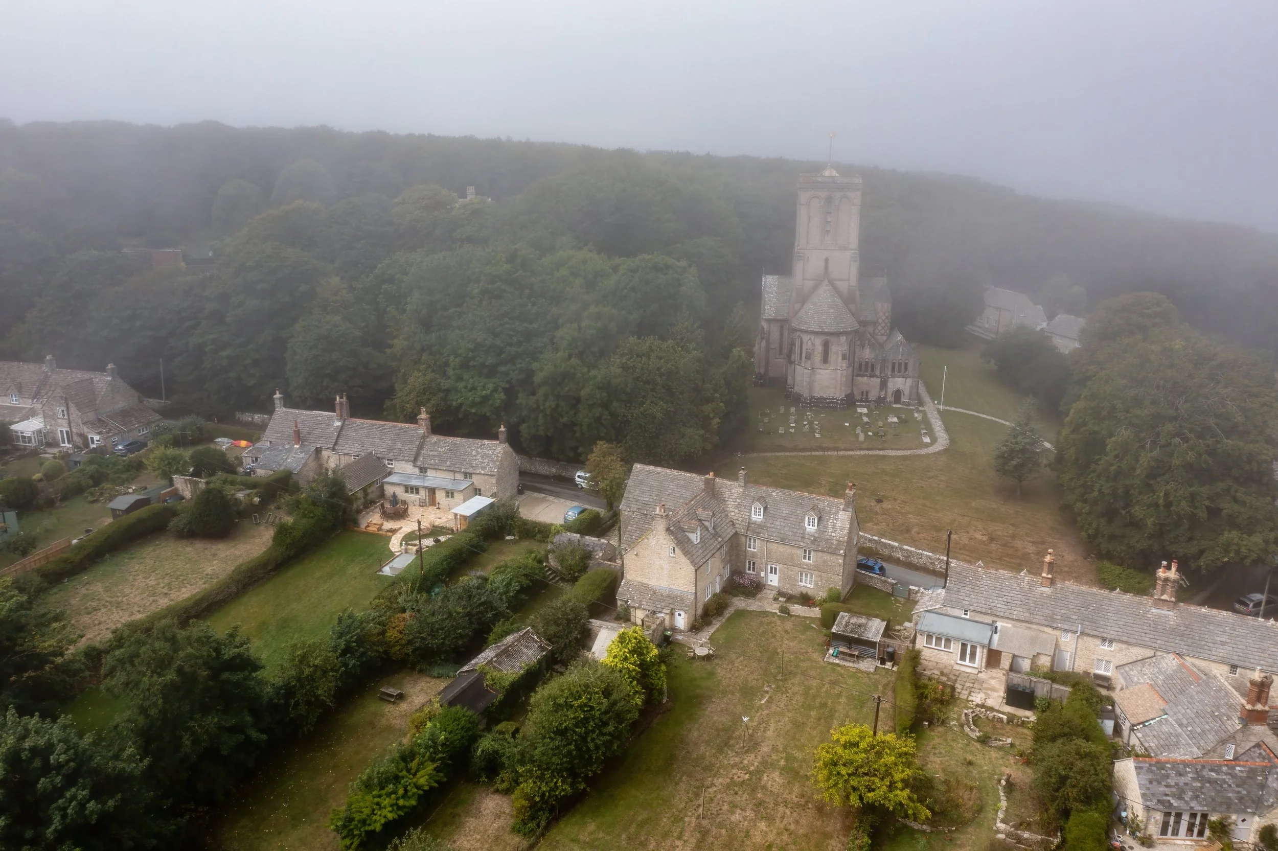 Aerial view of a small village with houses, gardens, and a large church on a hill, surrounded by trees and fog.
