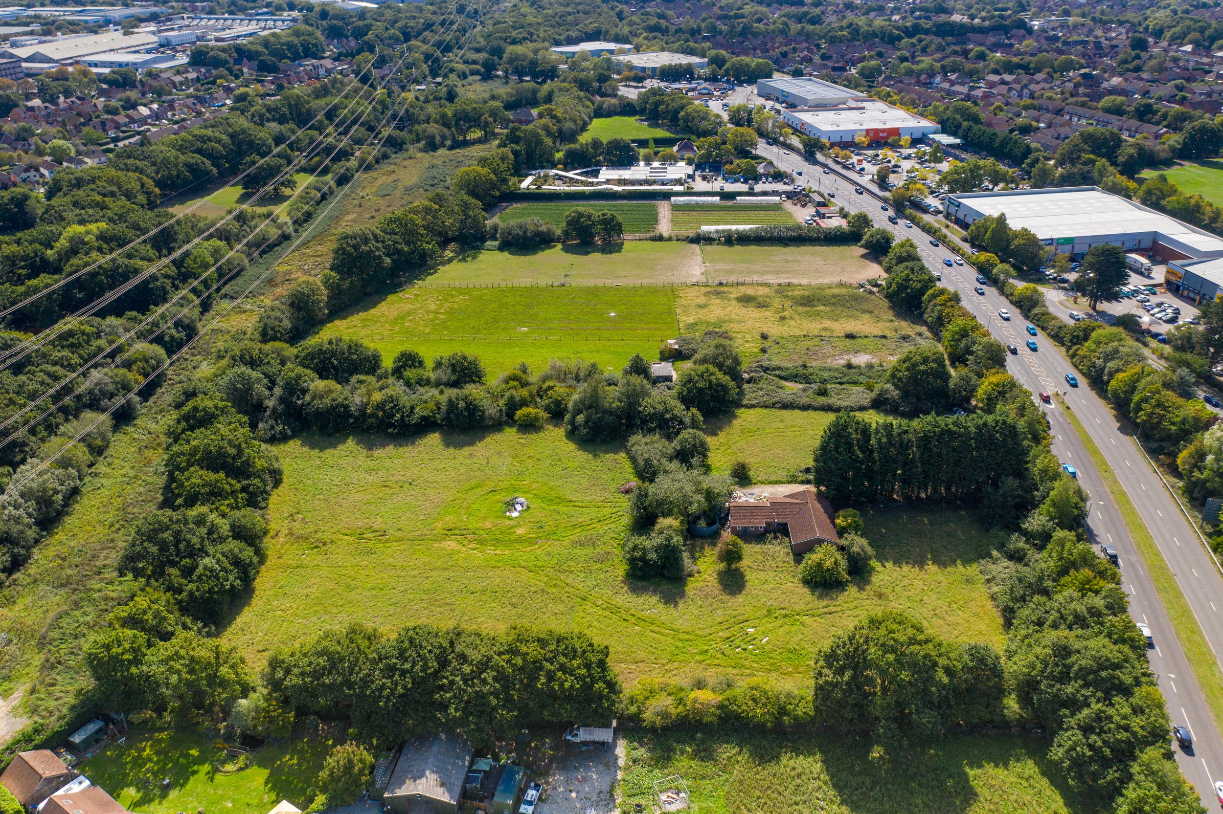 Aerial view of a suburban landscape showing a large green field with some trees, a house, and a road with cars parked and driving by. Residential houses and commercial buildings are visible in the background.