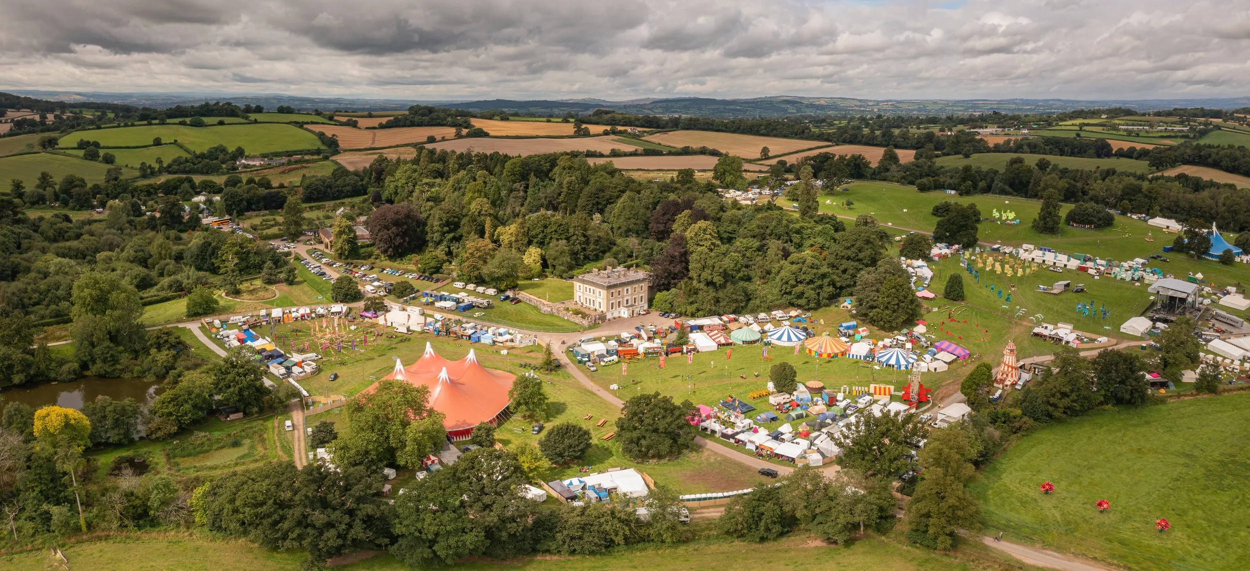 Aerial view of a fair with colorful tents, rides, and booths in a grassy park surrounded by trees and rolling hills.
