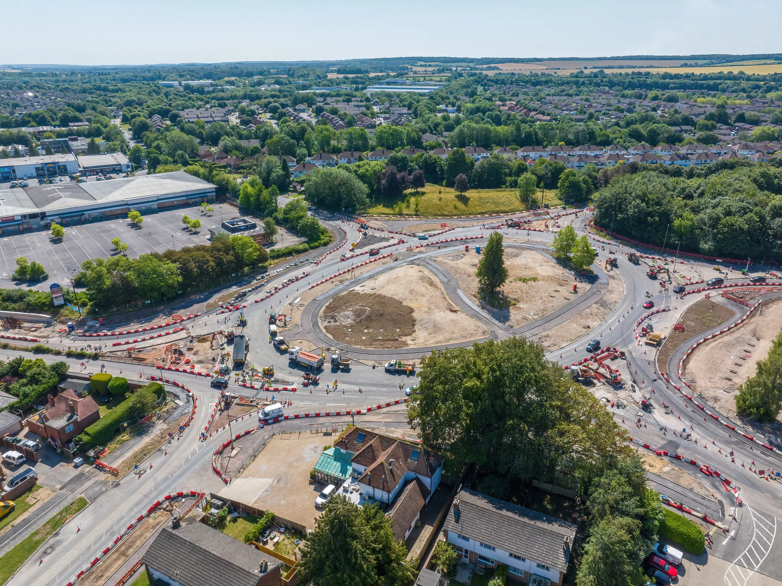 Aerial view of a roundabout under construction, with multiple roads, construction vehicles, orange and white safety barriers, residential houses, and green trees surrounding the area.