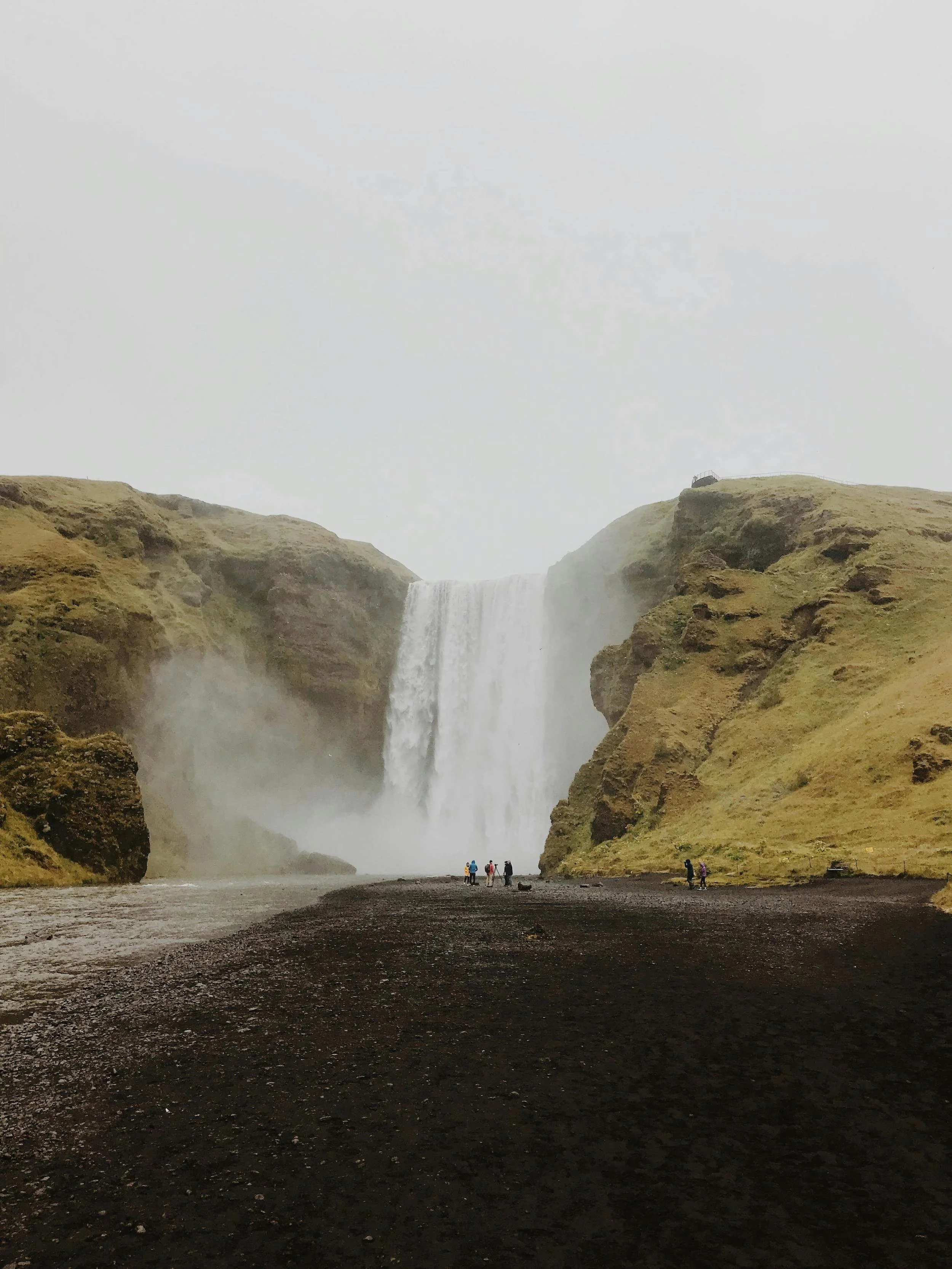 Waterfall cascading over a cliff with a misty backdrop, surrounded by mossy hills and a rocky riverbed. Small figures stand near the base of the waterfall.