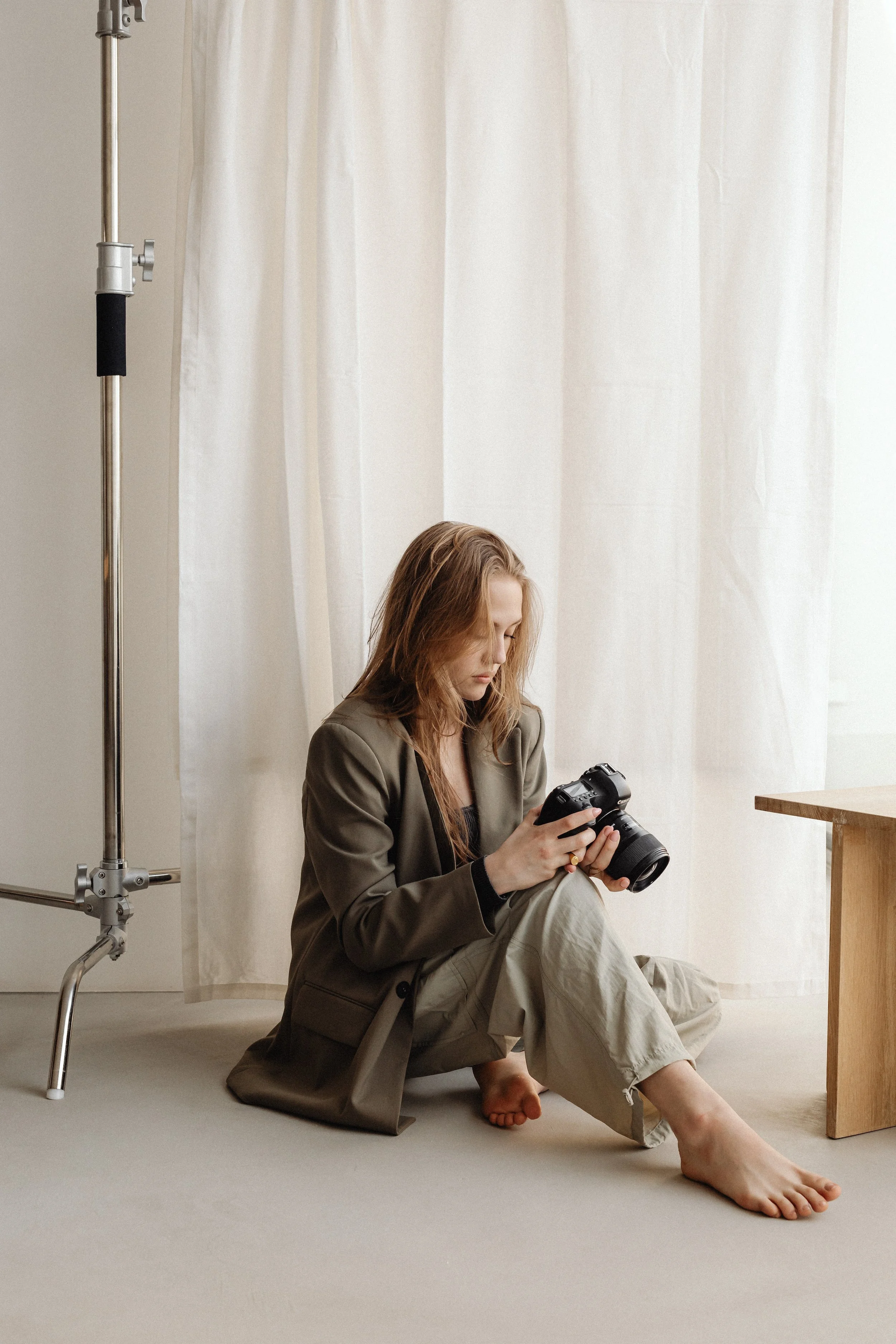 Person sitting on the floor holding a camera, wearing a blazer and light pants, with a curtain backdrop and a studio light stand nearby.