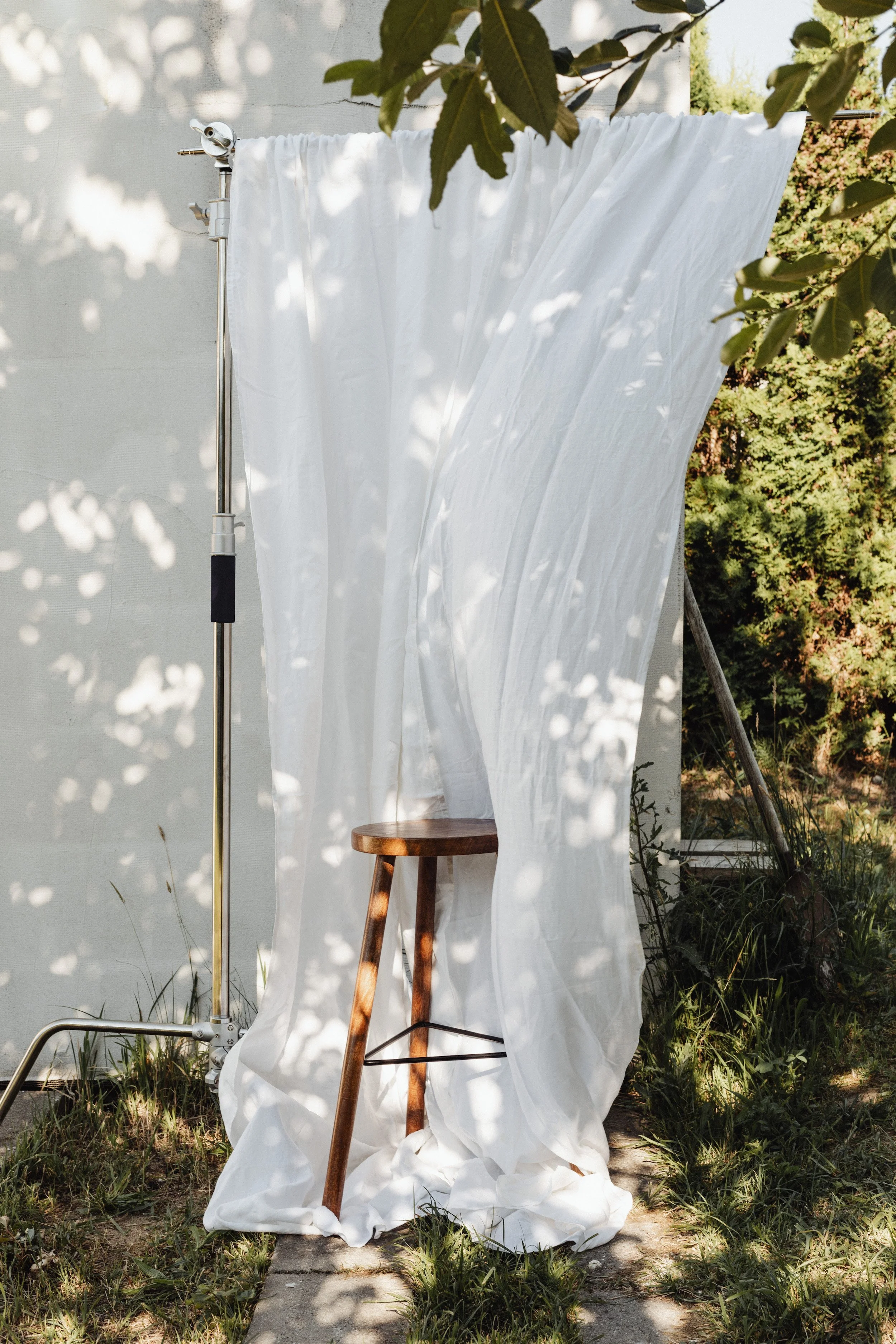 A wooden stool in front of a white curtain draped over a metal stand, outdoors with sunlight and shadows from leaves.