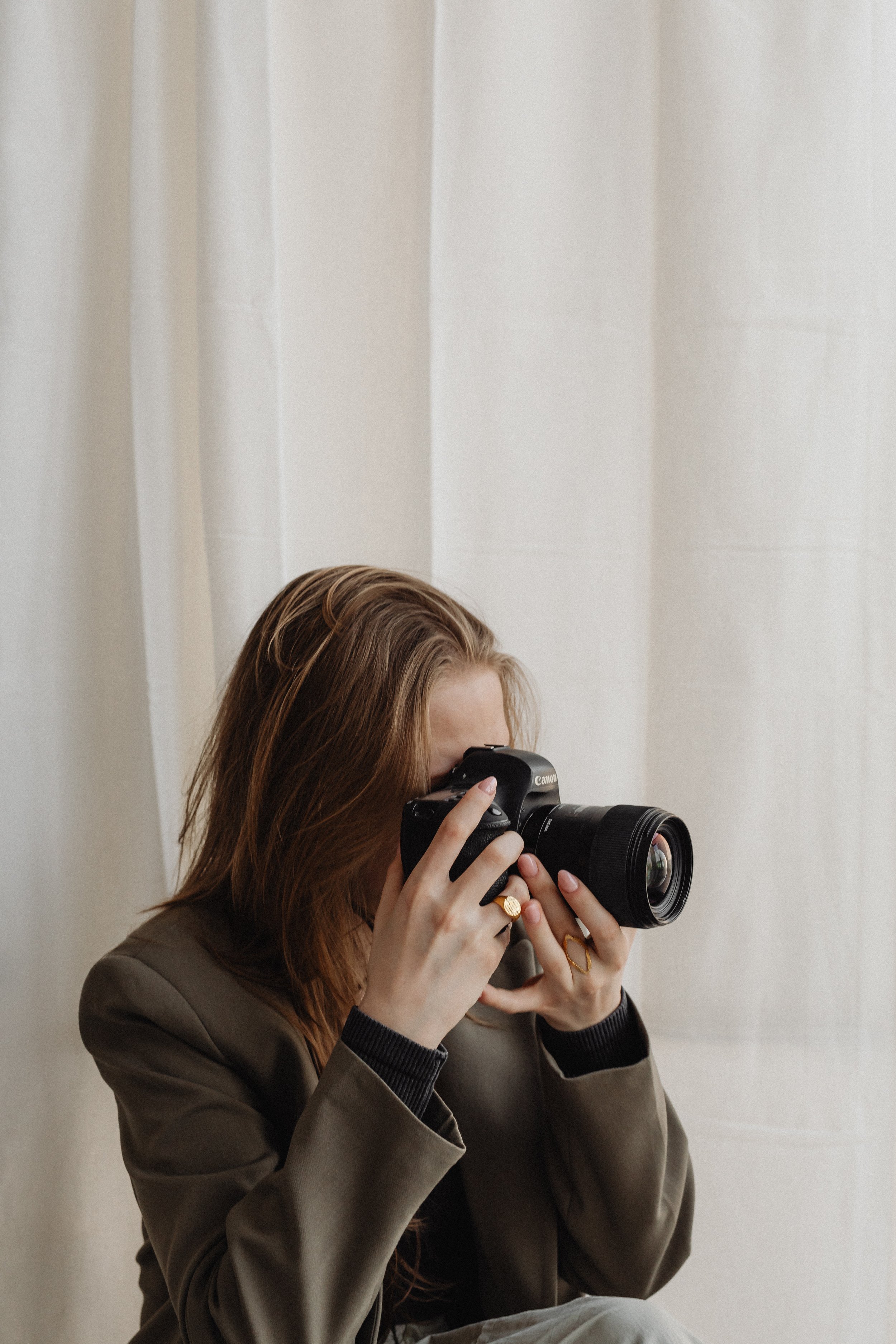 Person holding a camera, wearing a dark blazer, with a beige curtain background.
