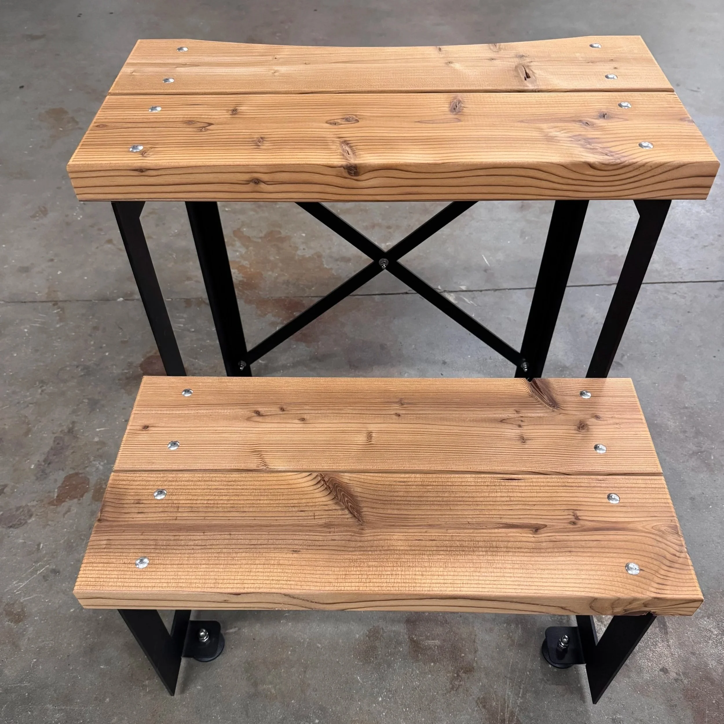 Een houten bank met metalen schroeven en zwarte metalen poten, geplaatst op een betonnen vloer.