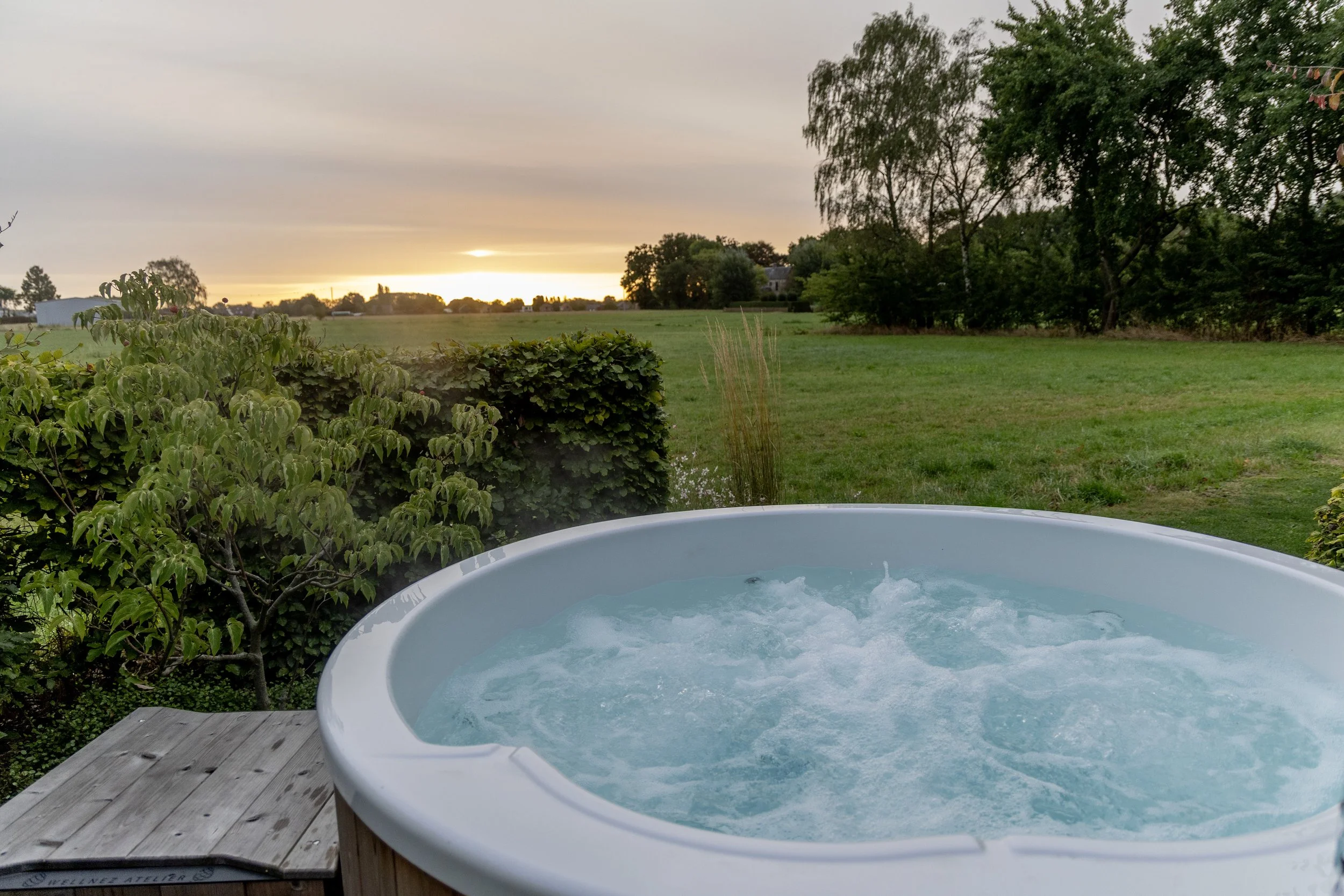 Wellnez Atelier  hottub gevuld met water en jets, omgeven door groen gras en bomen bij zonsondergang.