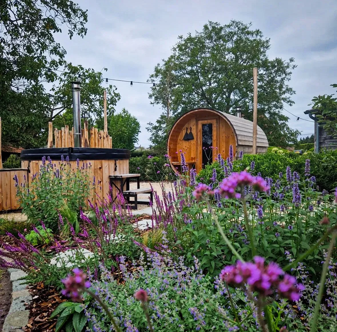 Een tuin met een hottub gebouwd door wellnez atelier, een houten tuinhut en een bloemenbed met purple en roze bloemen, omgeven door groen en bomen.