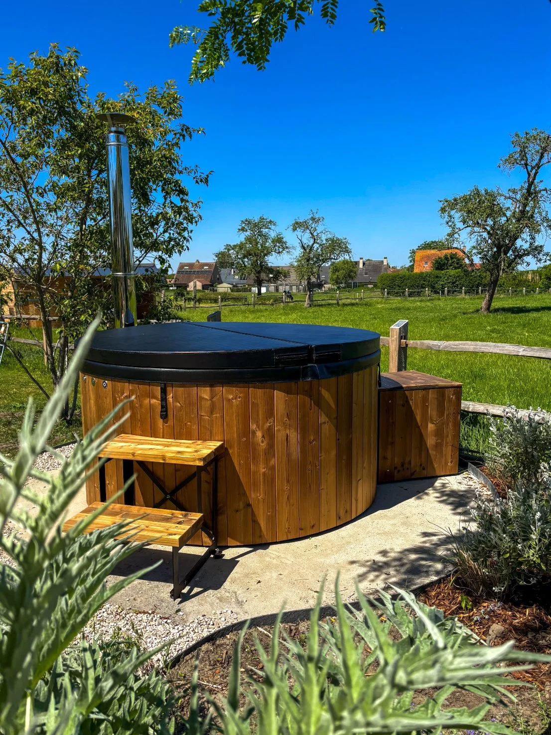Wellnez atelier Ronde houten hot tub met deksel, geïnstalleerd op een betonnen plaat in een tuin met groene grasvelden, bomen en huizen op de achtergrond onder een blauwe hemel  hottub op maat in tuin tevreden klant