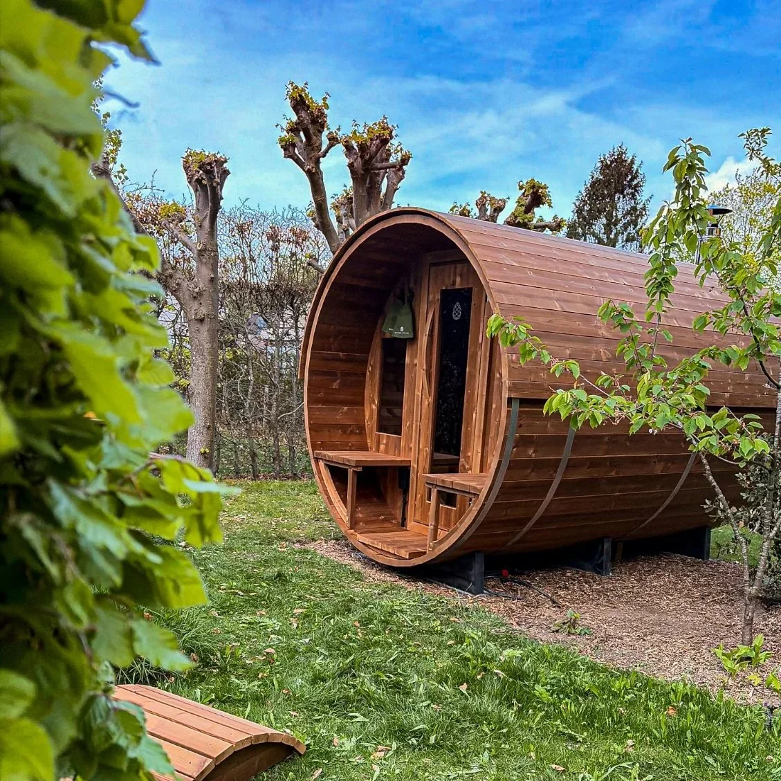 Handgemaakte barrelsauna gebouwd door Wellnez Atelier in een tuin in Gent met gras, bomen en een blauwe hemel.