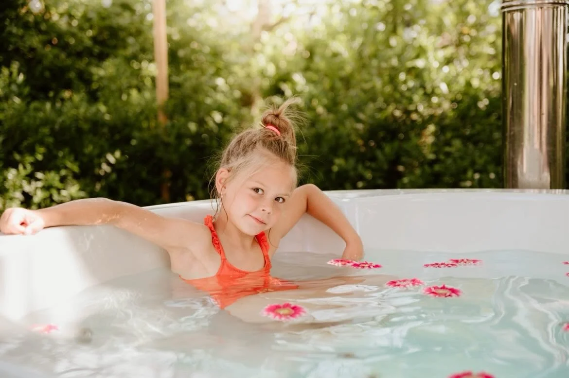 Een meisje in een oranje zwemoutfit zit in een hottub van Wellnez Atelier met roze bloemen op het water, omringd door groene struiken.