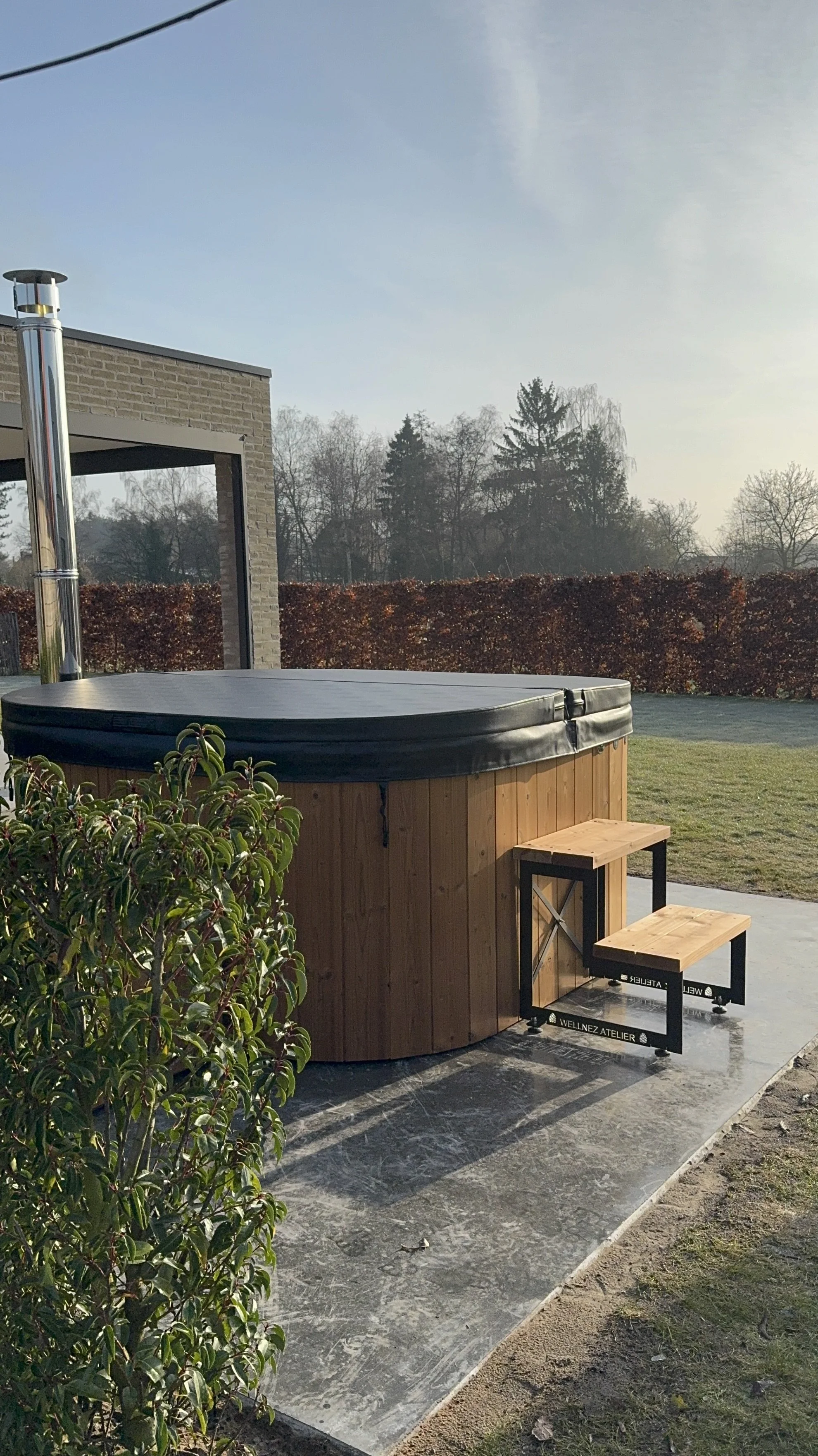 Een houten hot tub gebouwd door Wellnez Atelier met een beschermkap, naast een tuin en een grasveld. 