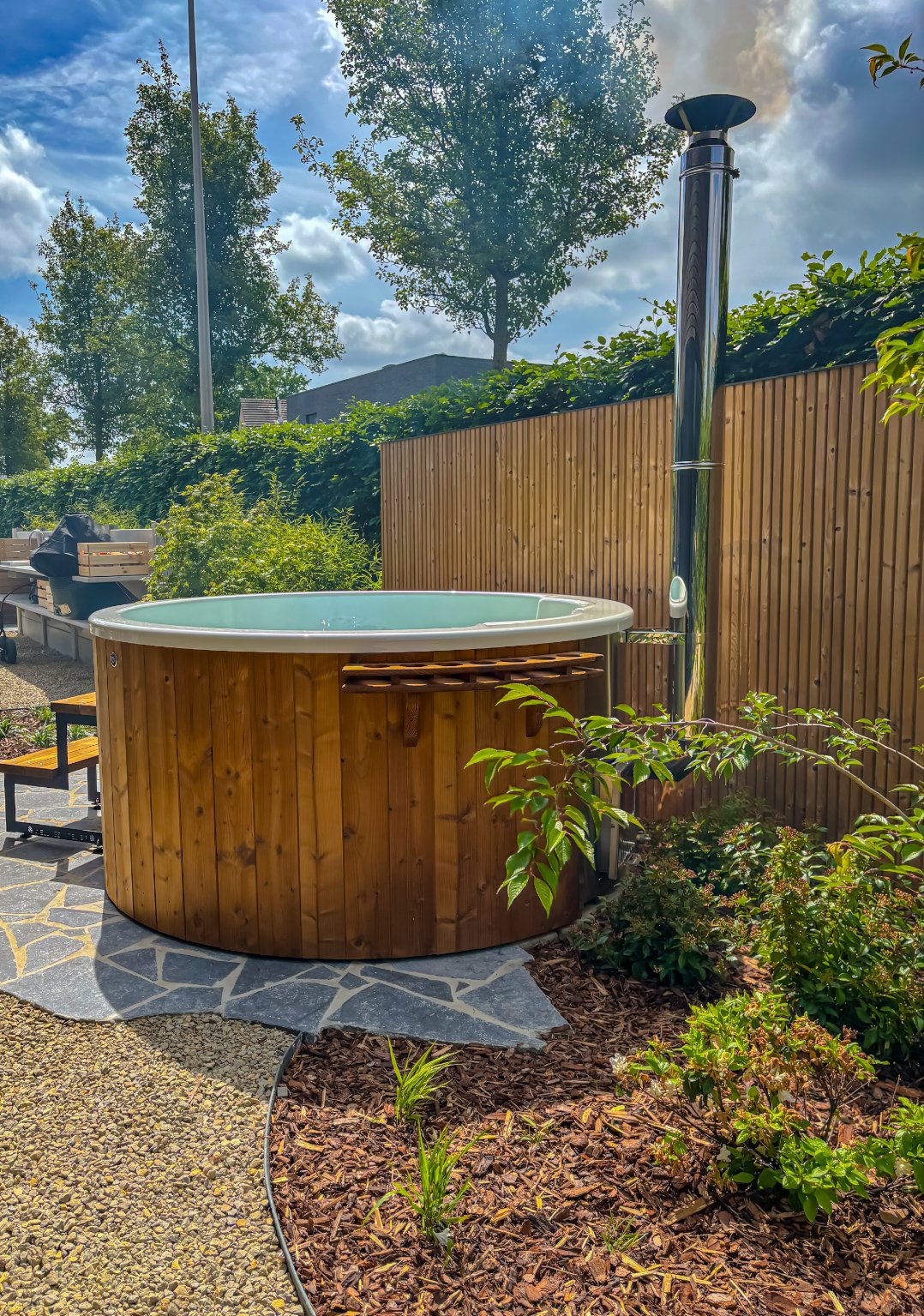 Hottub op maat met thermo vuren gebouwd door Wellnez Atelier, omgeven door tuin met planten en bloemen, houten schutting en helder blauwe hemel met wolken