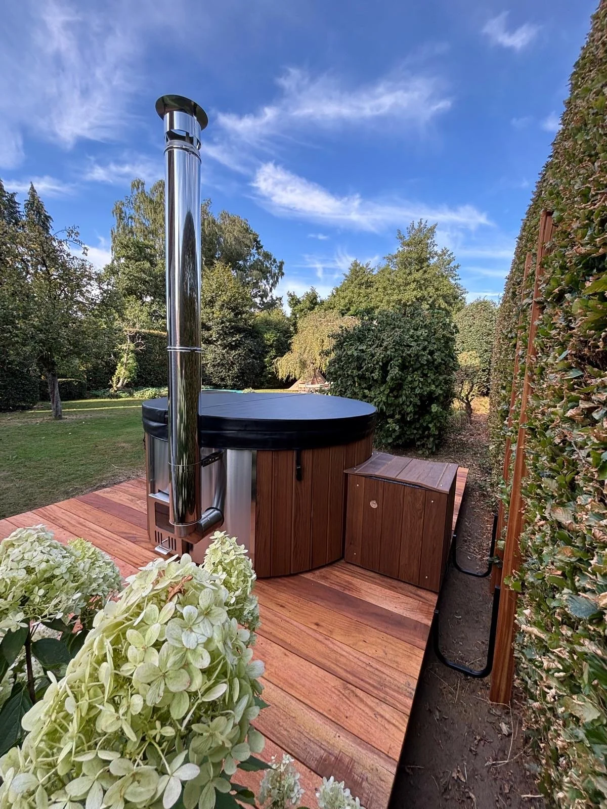 Een houten terras met een houten hottub met een zwart deksel gebouwd door Wellnez Atelier, omlijst door groene planten en bomen onder een blauwe hemel.