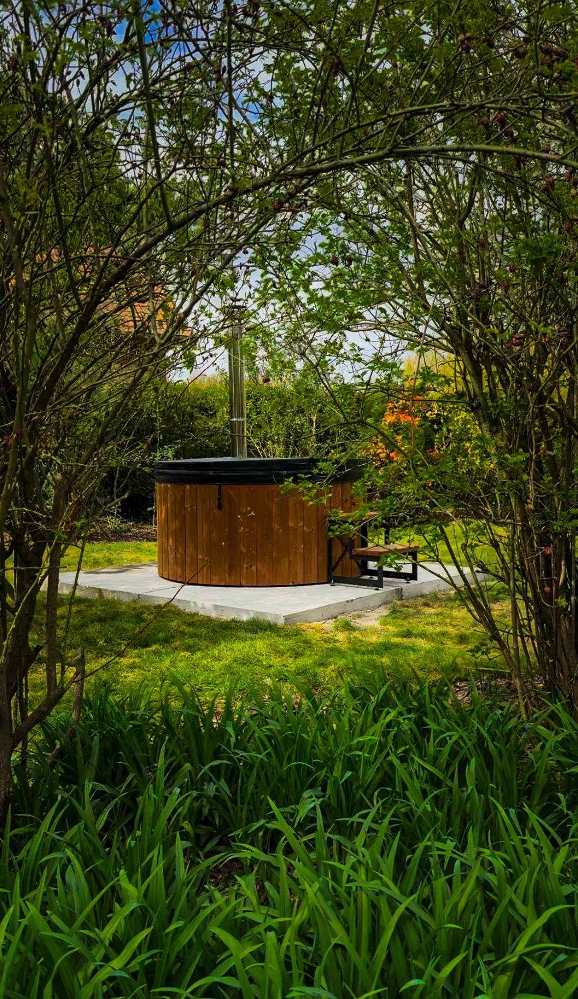 Een houten hottub gebouwd door wellnez atelier in een tuin, omgeven door groen gebladerte en bomen.