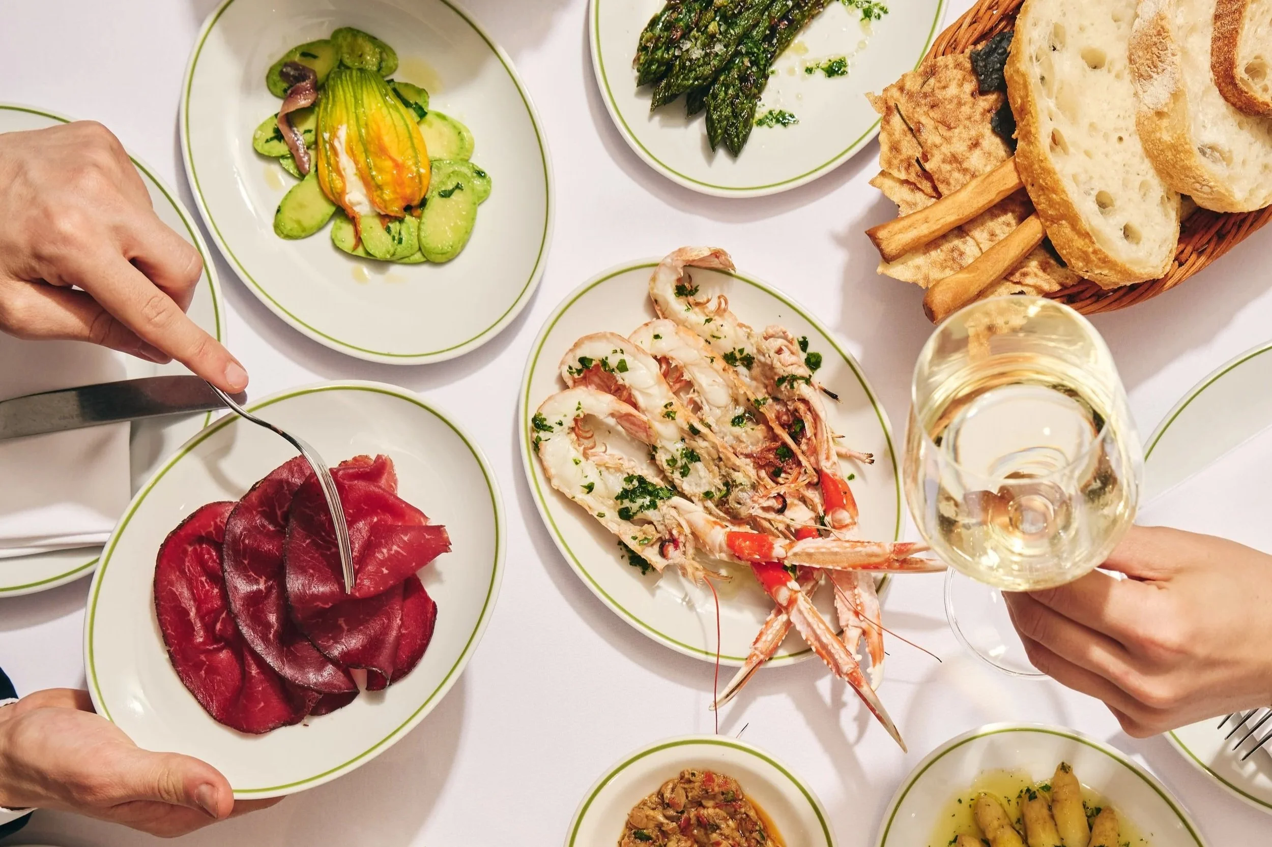 A variety of dishes on white plates, including sliced cured meat, a seafood dish with lobster, roasted asparagus, sliced cucumber salad, and a basket of bread with breadsticks. Someone is holding a glass of white wine.