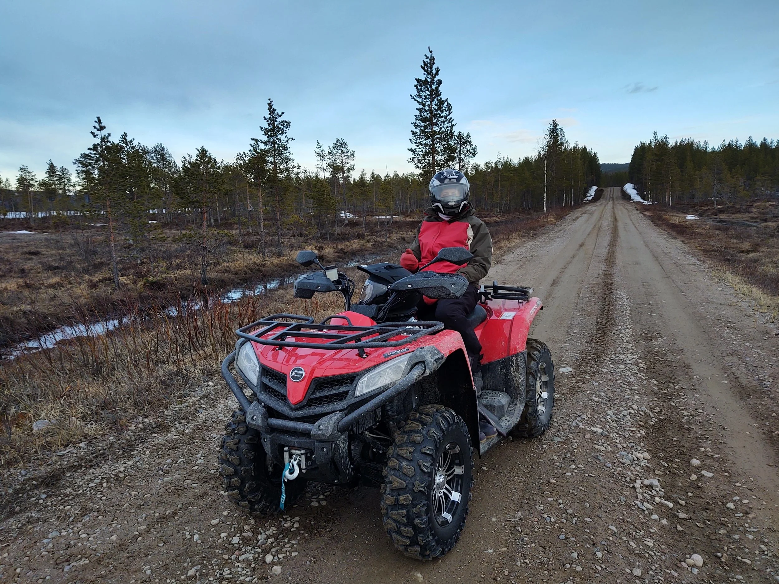 Person wearing a helmet and a red and brown jacket sitting on a red all-terrain vehicle (ATV / Quad) on a dirt road in a rural area with trees on both sides and patches of snow.