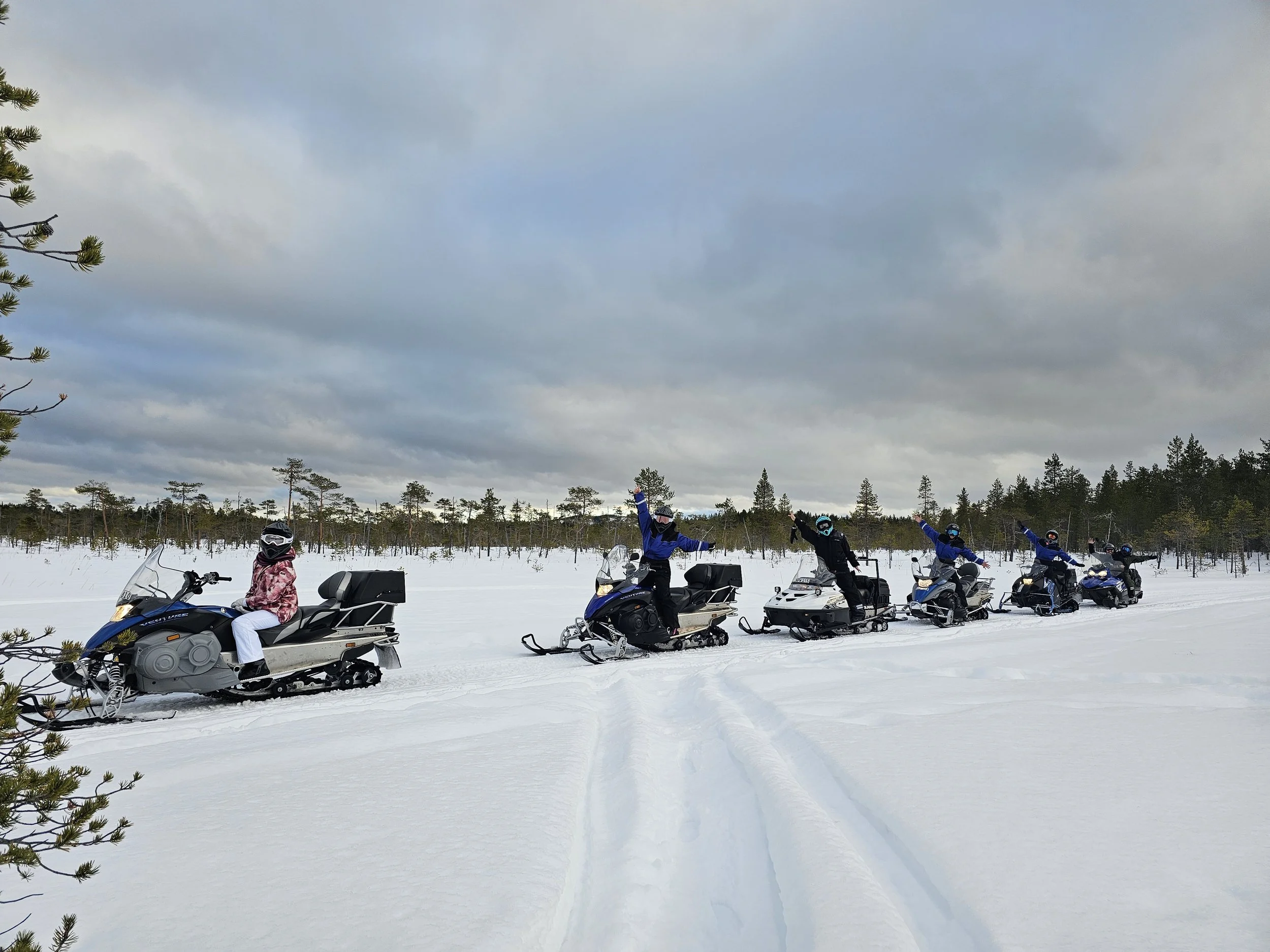 Group of six people on snowmobiles in a snowy landscape under cloudy sky, with trees in the background and one person in front wearing a pink jacket and white pants, others waving.