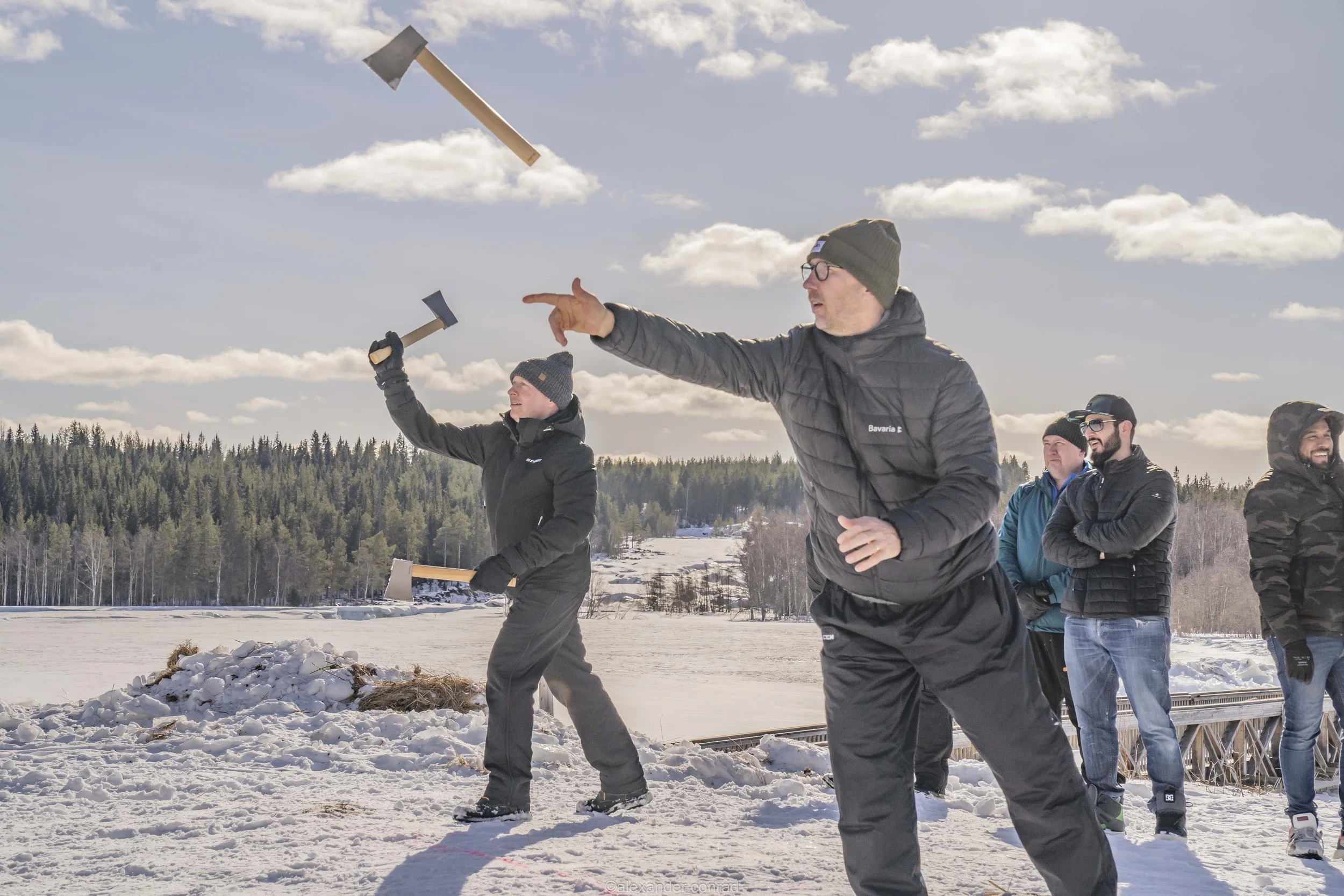 Group of people outdoors in a snowy landscape, some holding axes, with one person pointing towards the sky and another swinging an axe, on a sunny day with clouds and a forest in the background.