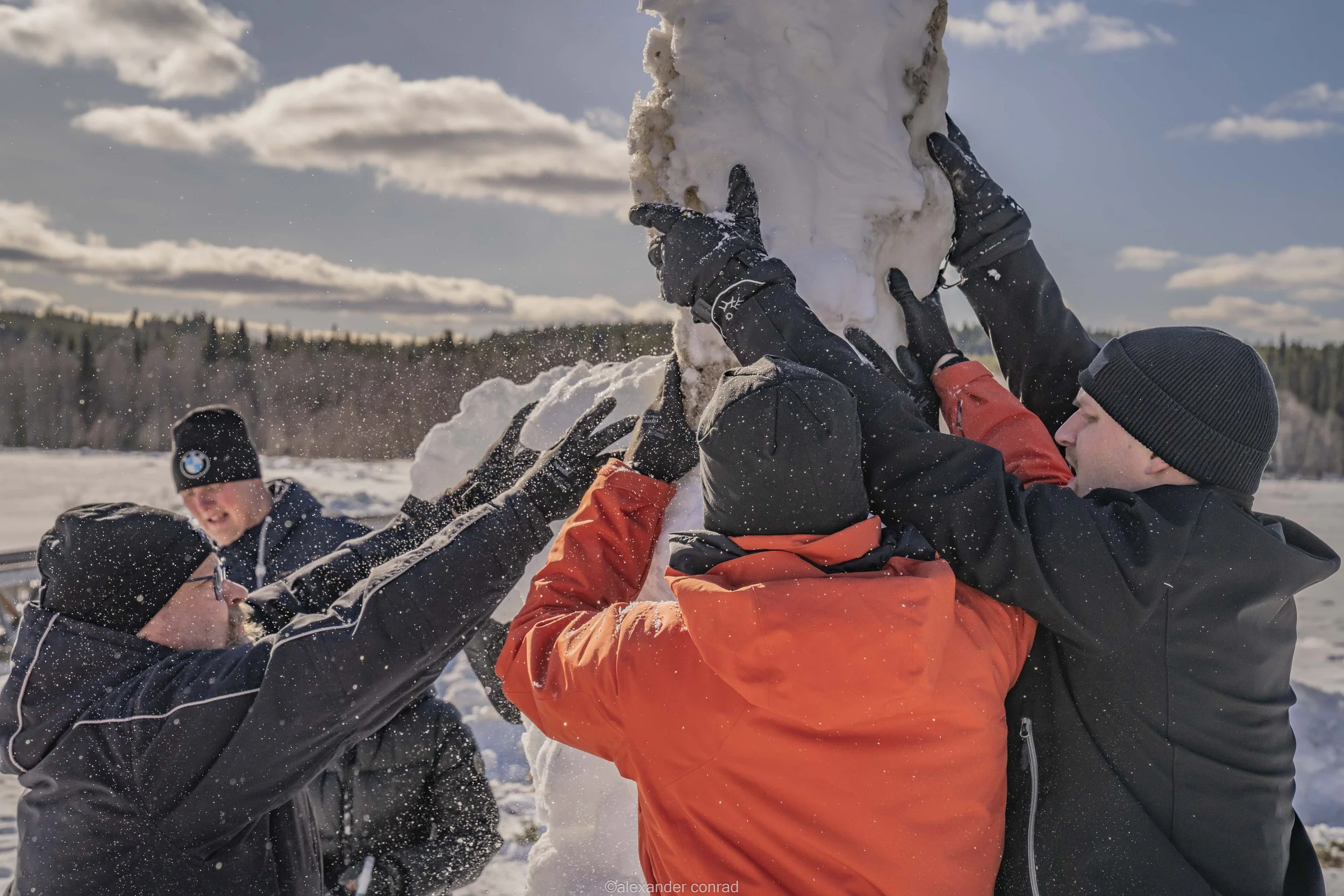 Group of people in winter clothing building a snowman outdoors in a snowy landscape, with trees and a partly cloudy sky in the background.
