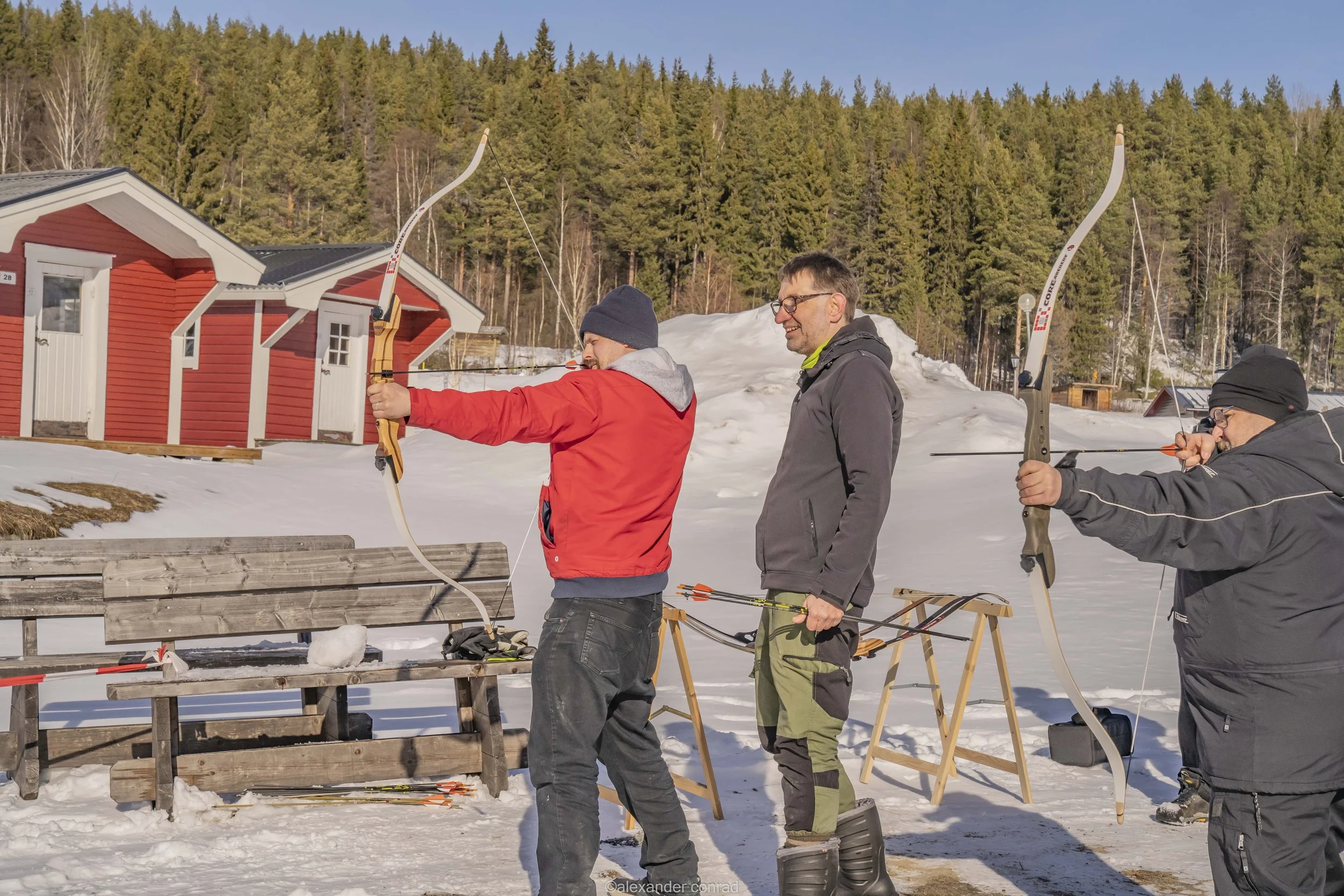 Three men practicing archery outdoors on a snow-covered area with red cabins and a pine forest in the background, under a clear blue sky.