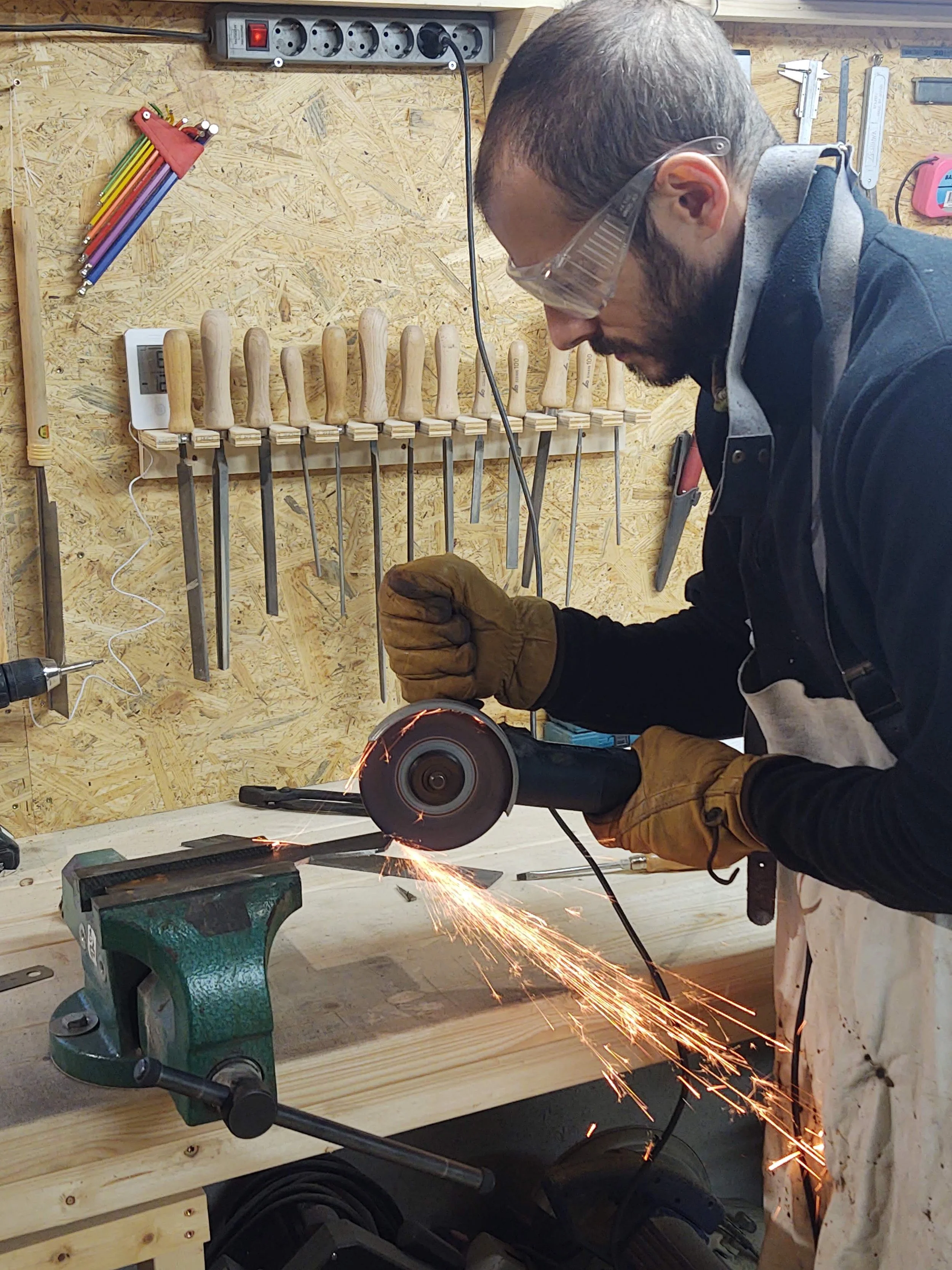 A man wearing safety glasses and gloves is cutting metal with an angle grinder, sparks flying in a woodworking workshop with hand tools on a wooden workbench and tools hanging on the plywood wall.