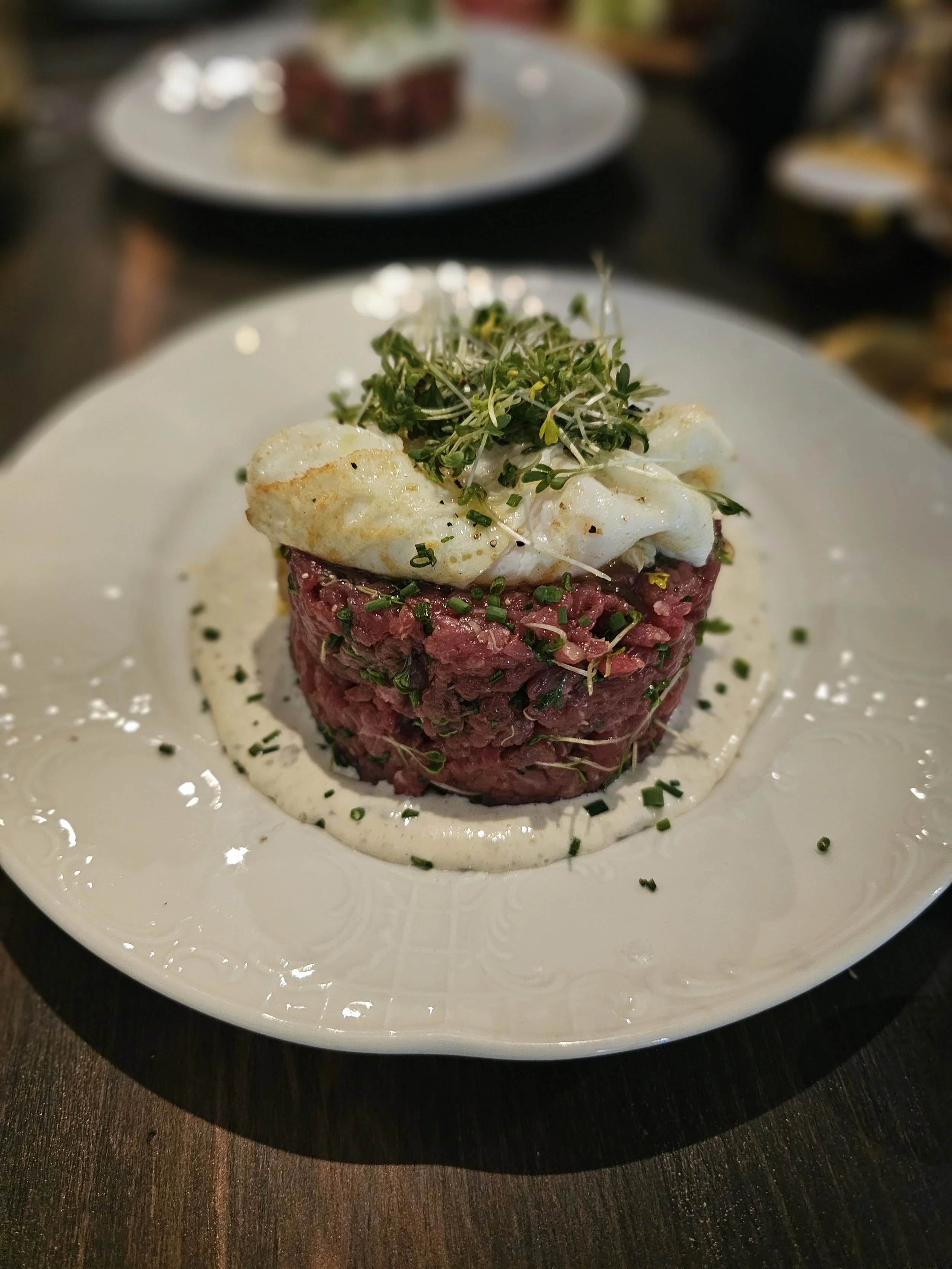 A plated beef tartare topped with a poached egg and microgreens, garnished with sauce on a white ornate plate.