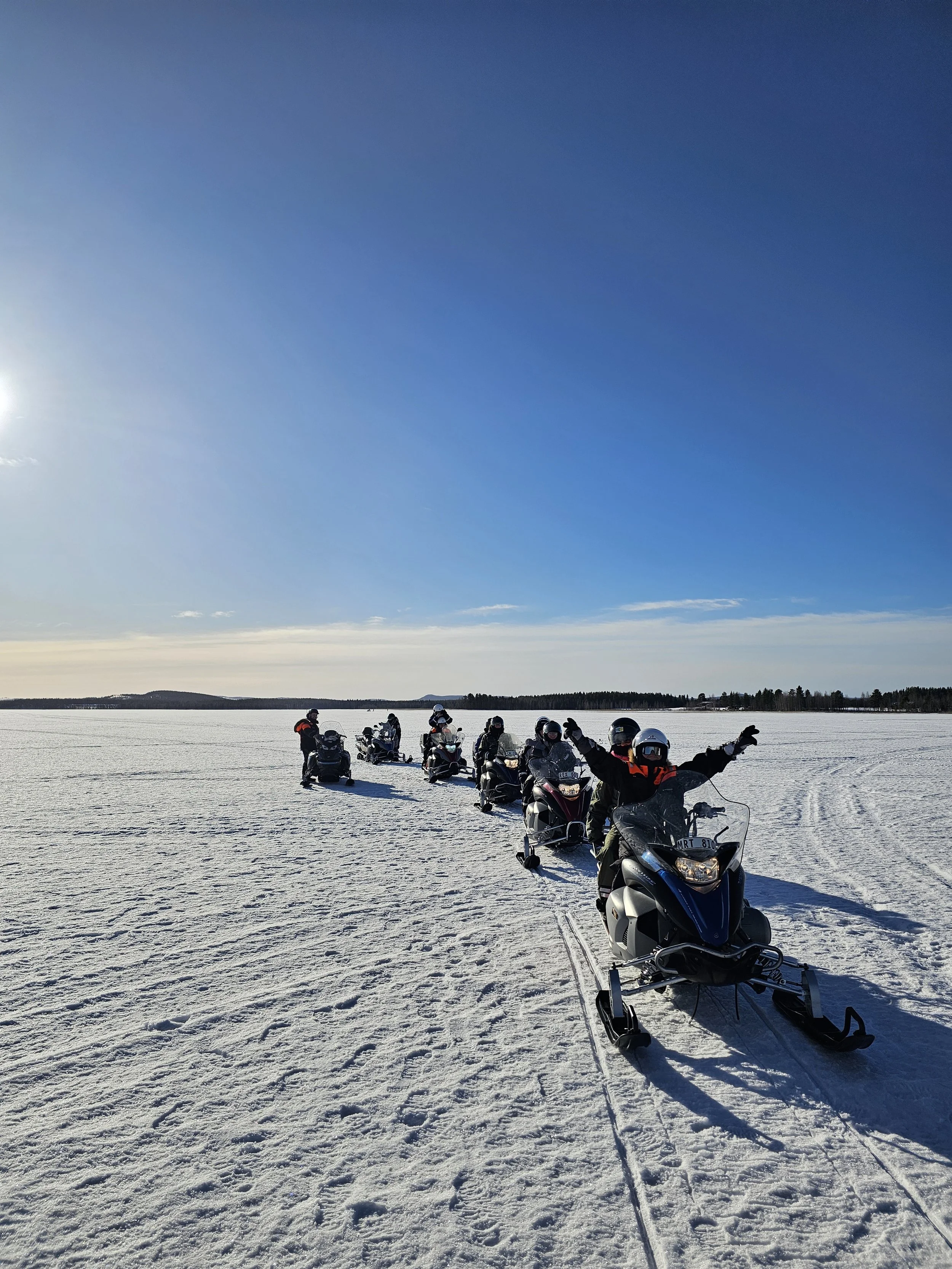 A group of people riding snowmobiles across a snowy landscape under a clear blue sky with the sun shining.