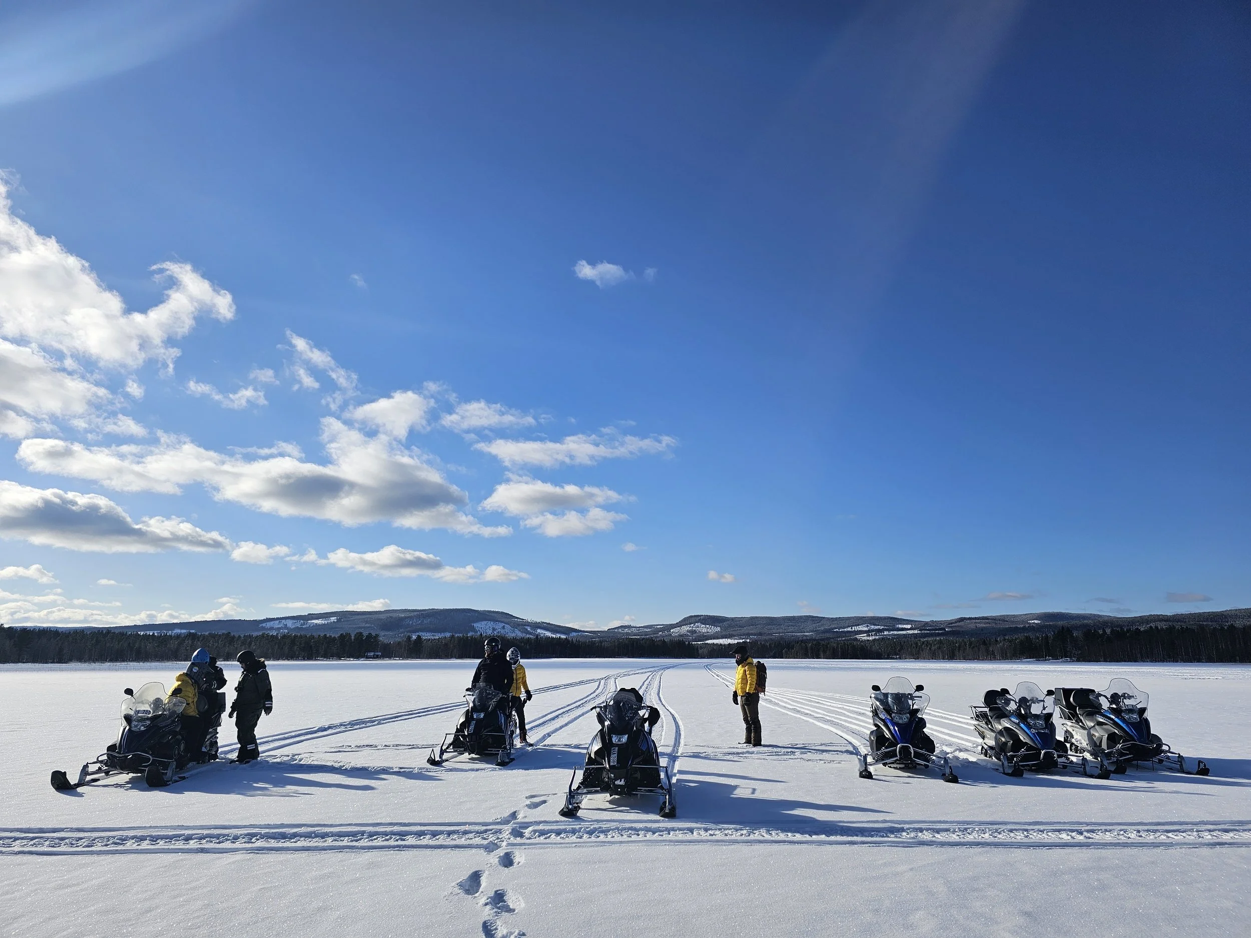 Group of people in winter gear with snowmobiles on a snow-covered lake under a blue sky with clouds.