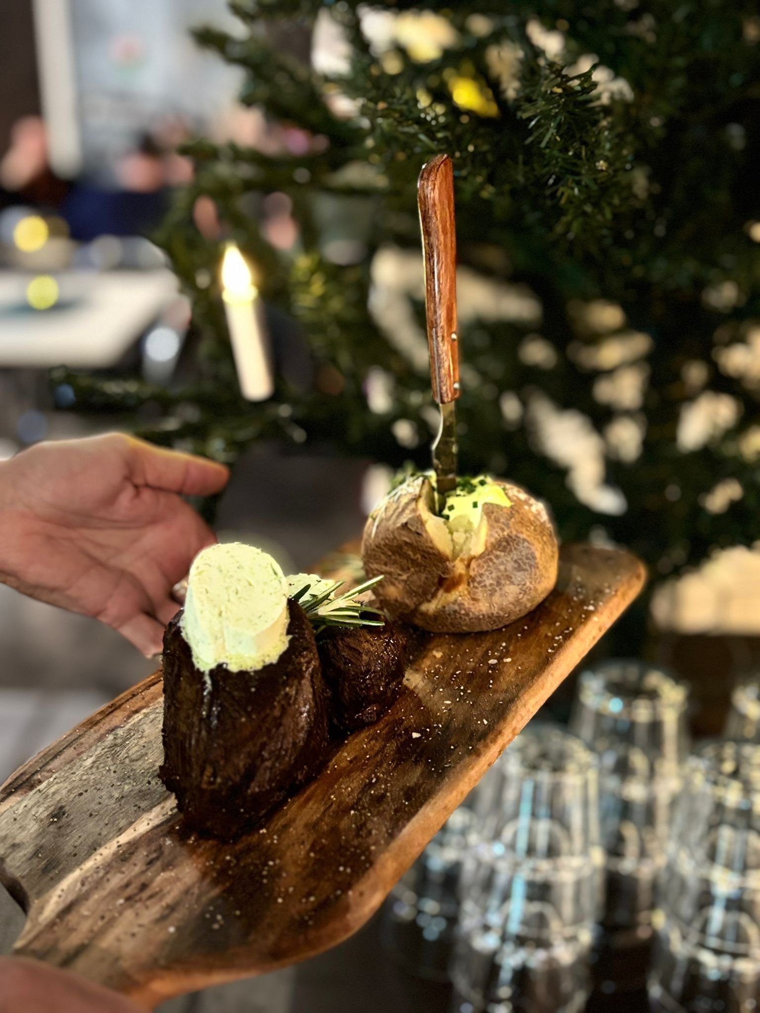 A wooden platter with two roasted potatoes topped with herb butter, garnished with garlic and fresh herbs, held over a blurred background of a Christmas tree and glasses.