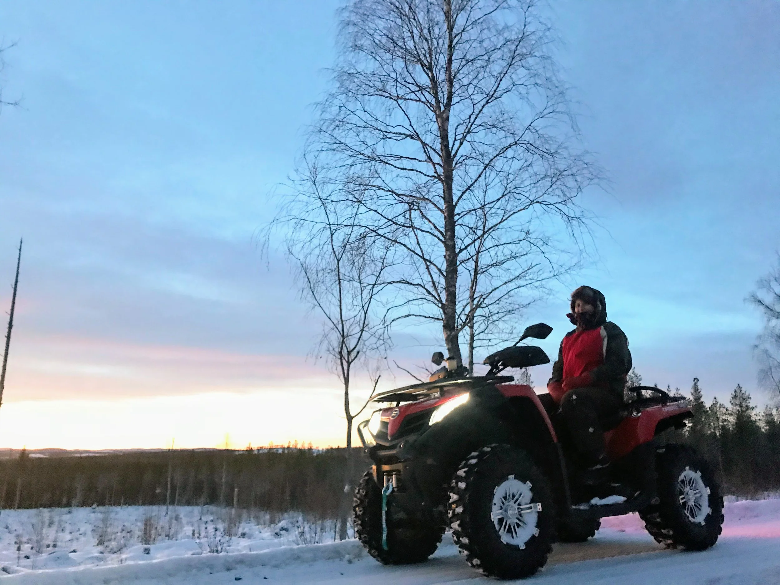 Person wearing a red jacket and helmet sitting on a black and red ATV / Quad on a snow-covered road with leafless trees and a sunset sky in the background.
