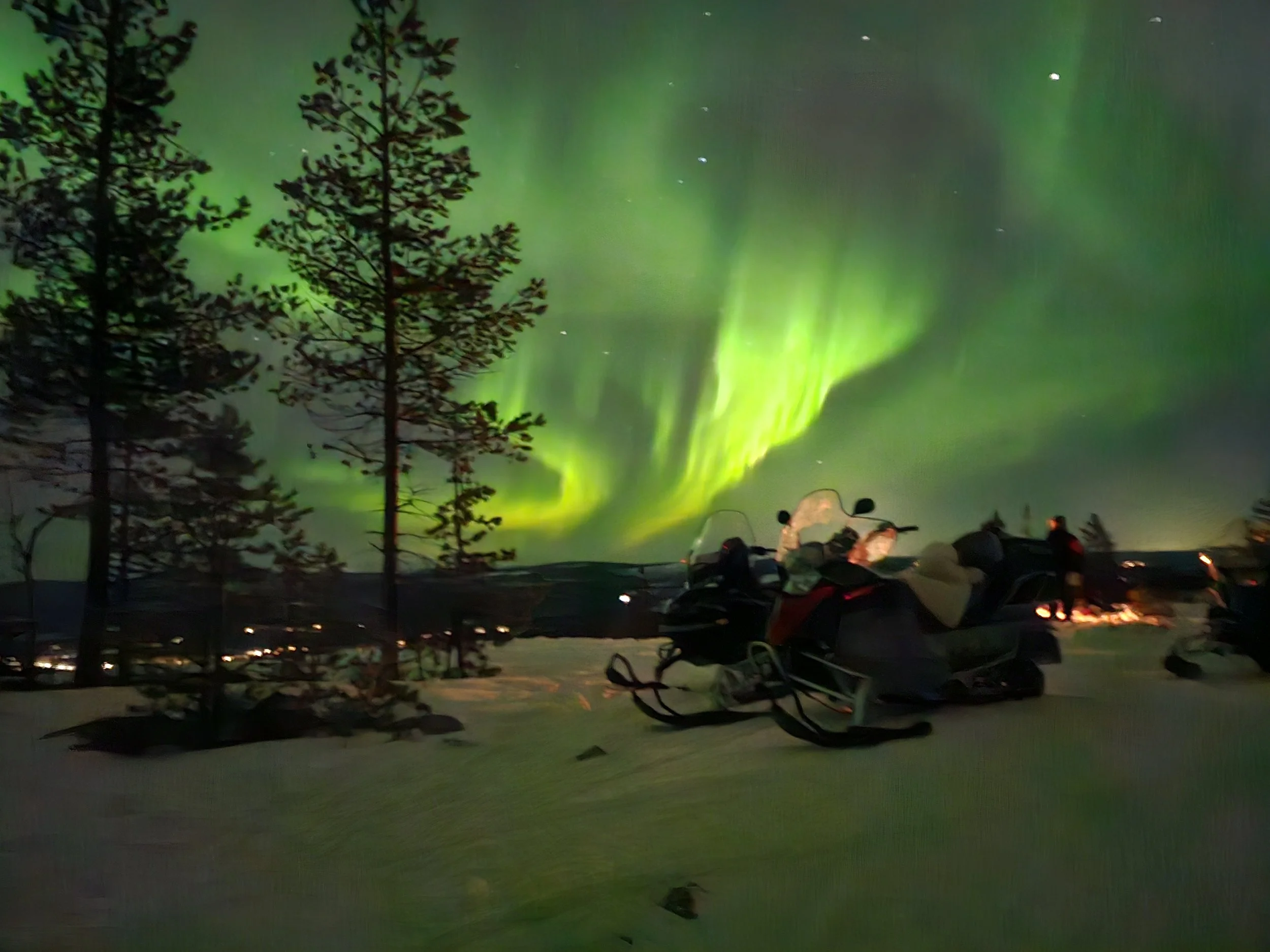Snowmobiles parked under the northern lights, aurora borealis, while guests warm around a fire