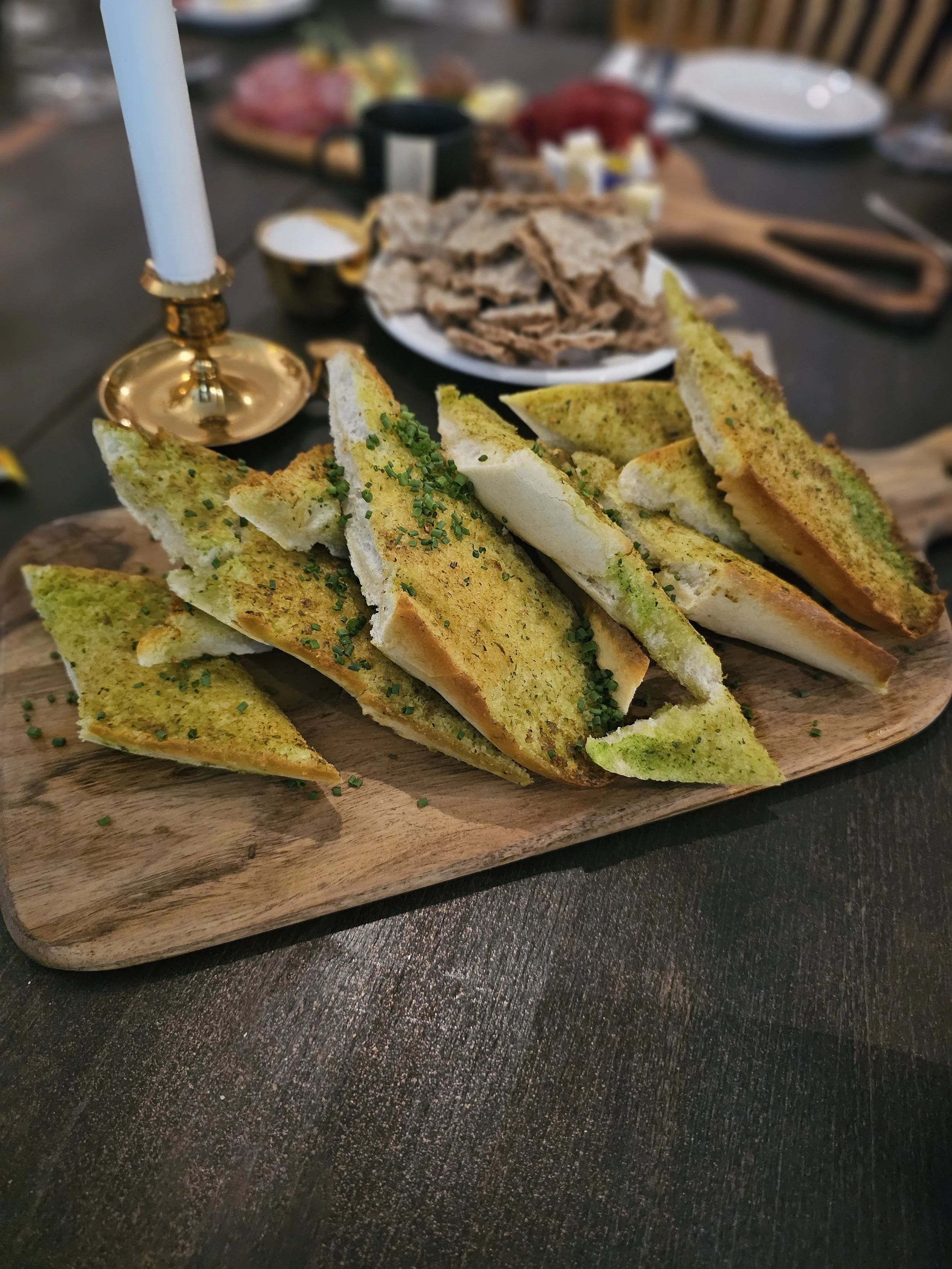 Close-up of garlic and herb bread slices garnished with chopped chives, served on a wooden board at a dinner party.