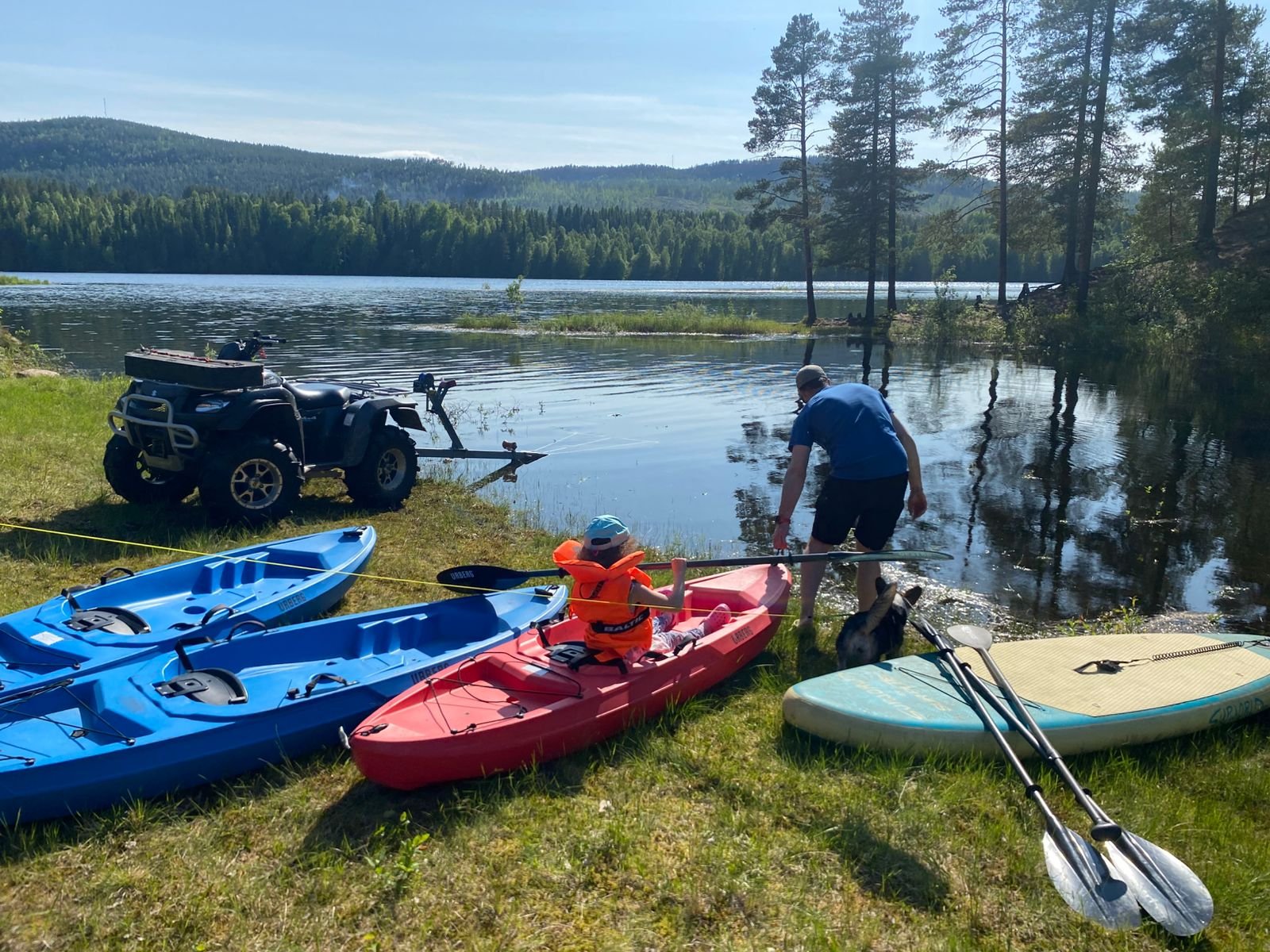 Two people and a dog preparing kayaks and paddleboards by the lakeside with an ATV / Quad on a sunny day, with a background of trees and mountain.