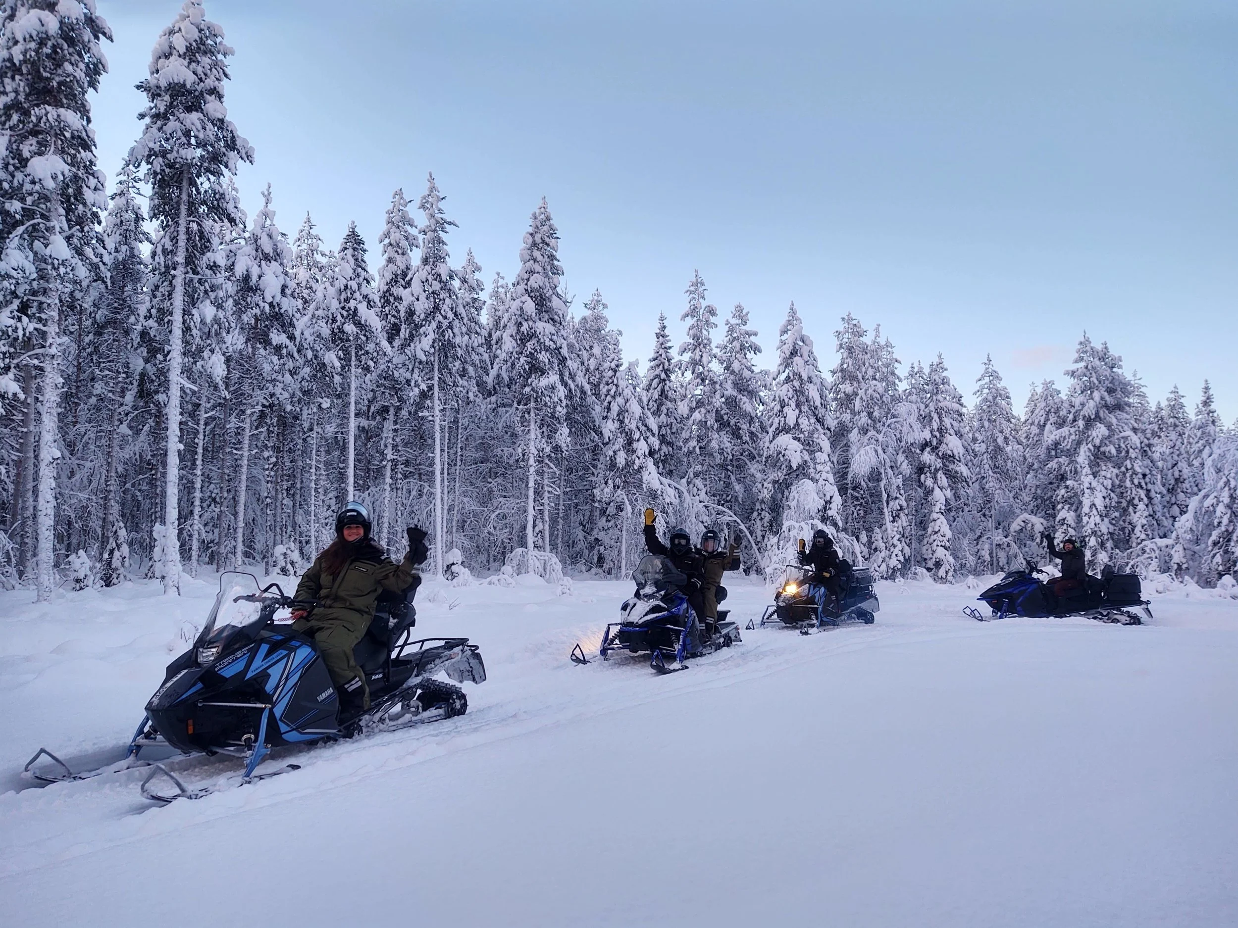 Guests on snowmobile tour in a snowy, forested area with tall, snow-covered trees and a clear sky.