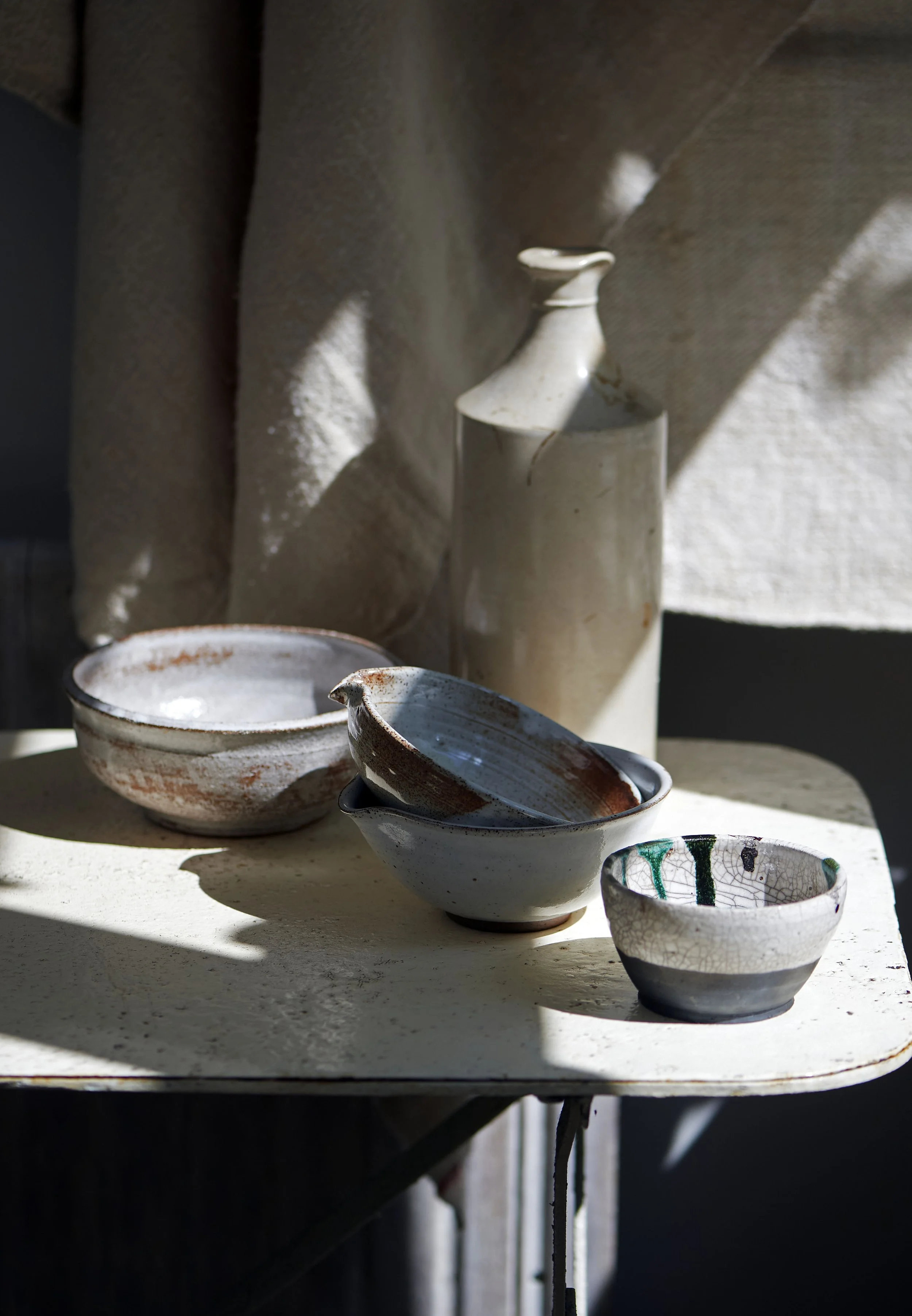 Several handmade ceramic bowls and a ceramic pitcher resting on a white table, with natural light and shadows creating a warm atmosphere.