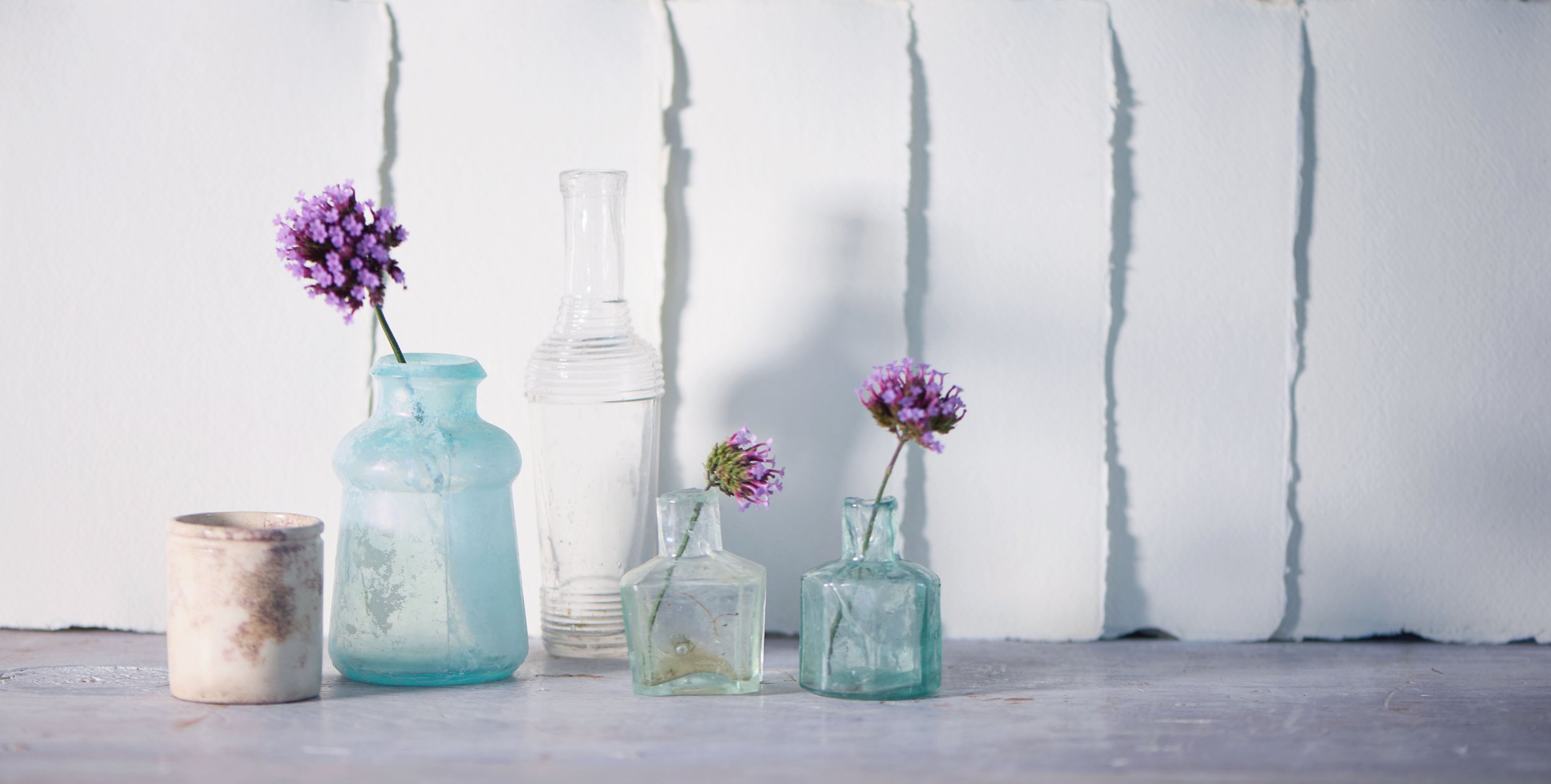 Four small glass bottles with purple flowers, a white ceramic cup, and a clear glass bottle on a light-colored surface with a white wall background.