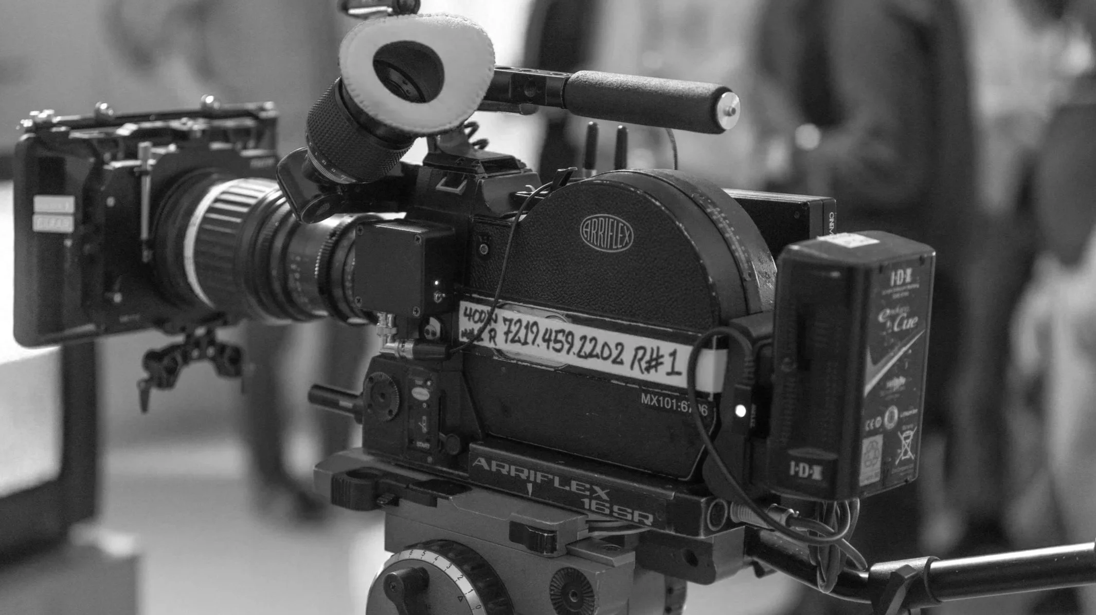 Film camera on a tripod with a red cable and a red label, in a setting with people in the background.