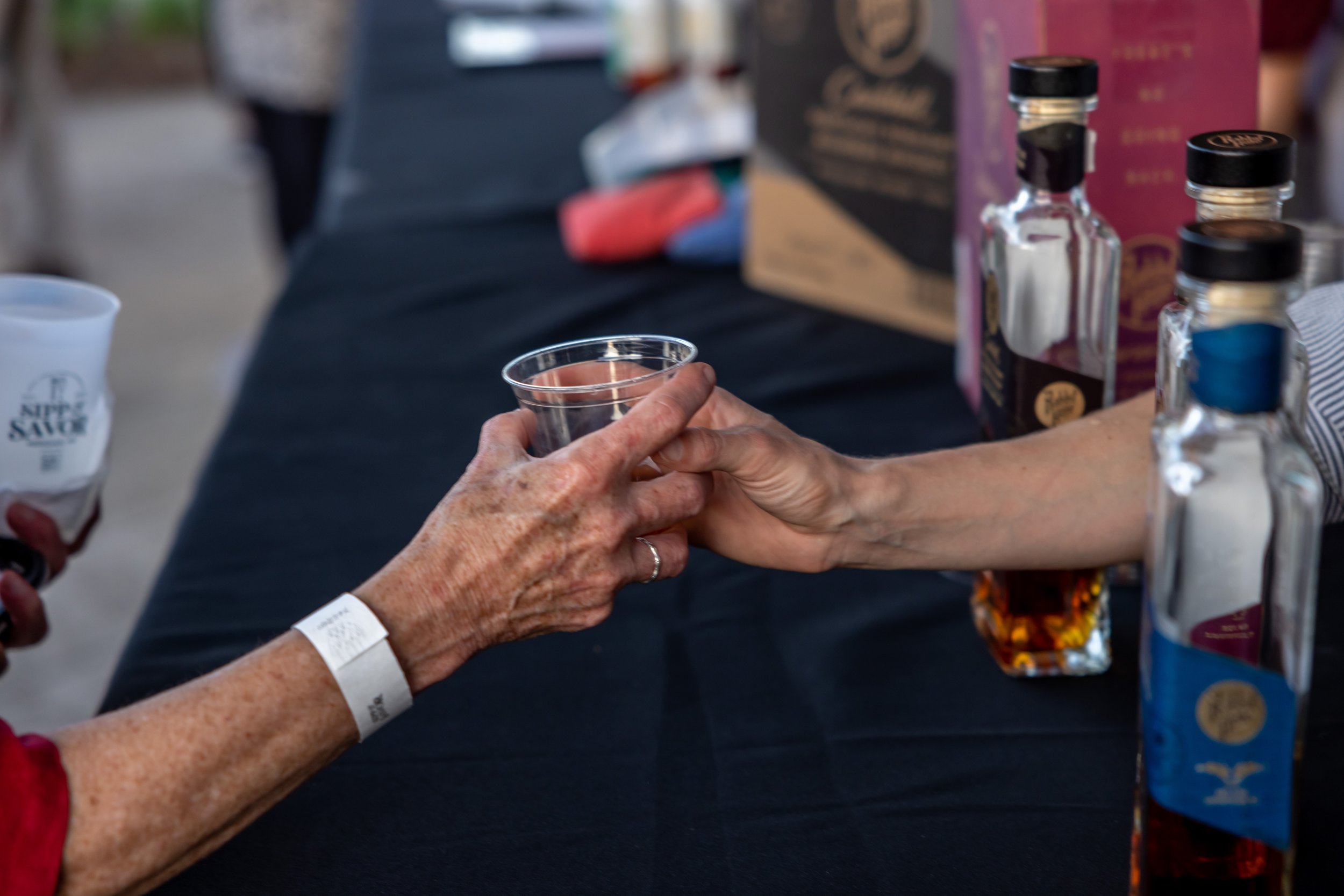 Two people exchanging a plastic cup at a table with bottles of liquor.