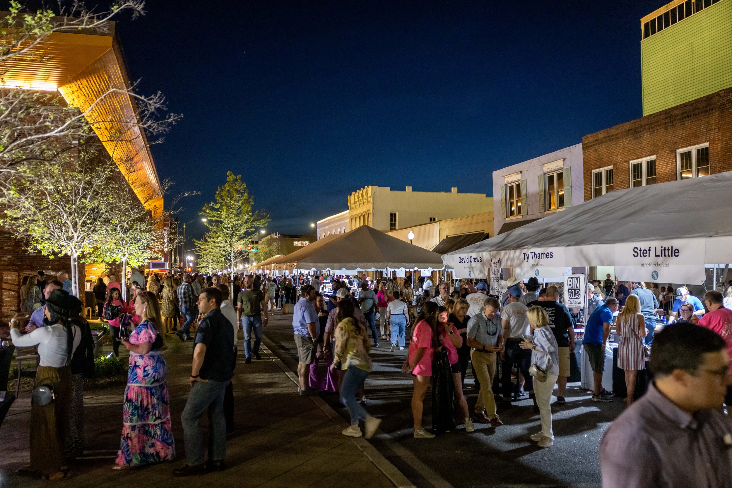 Outdoor evening street event with people gathered under tents, engaging in social activities. Buildings and trees are visible, with a colorful sky in the background.