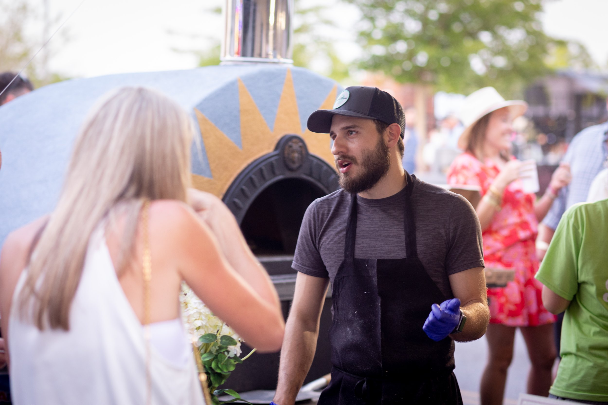 Outdoor event with a man wearing a black cap and apron talking to a woman. A wood-fired oven is in the background, and people socialize nearby.