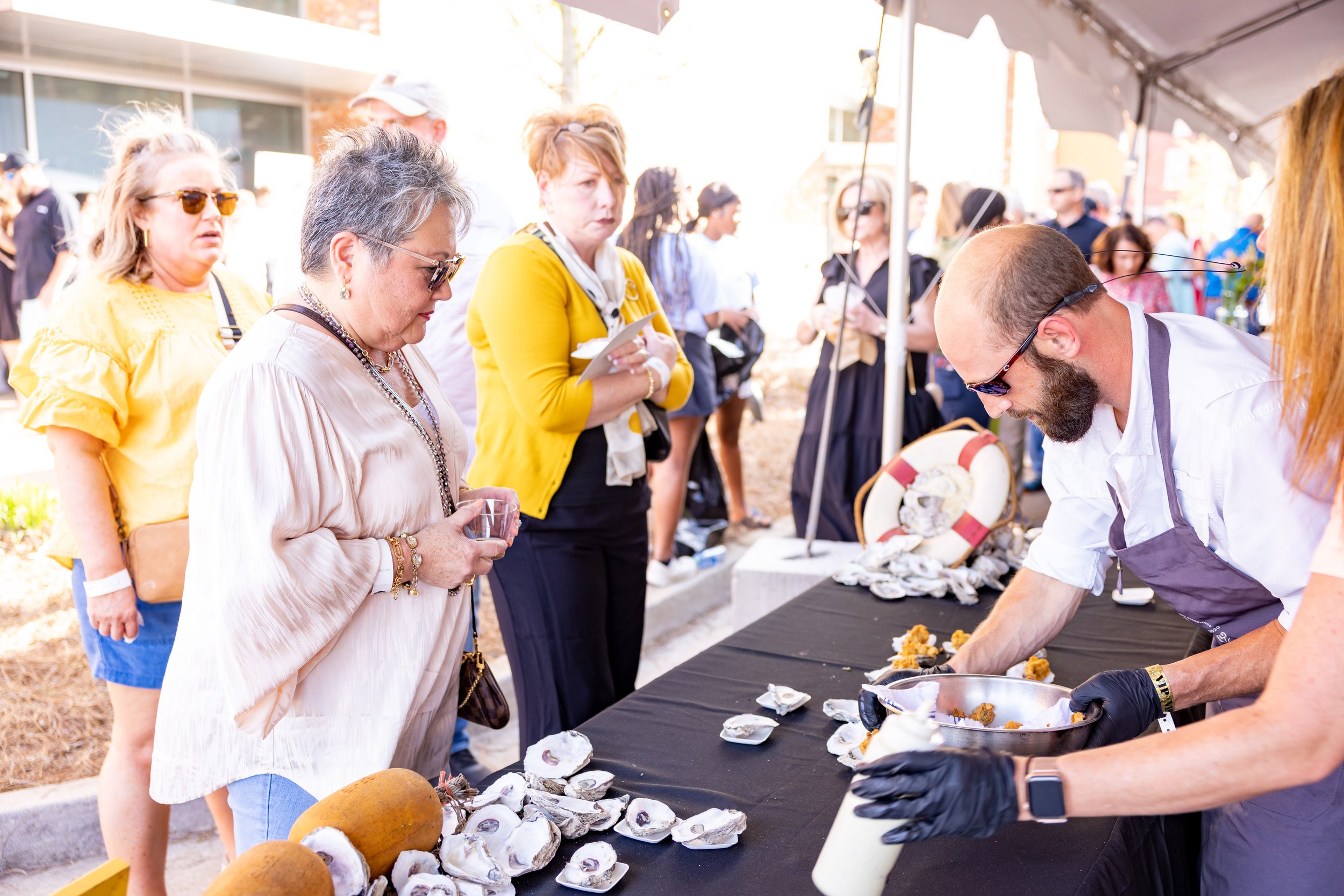 People gathered at an outdoor event, watching a man in a white shirt and apron preparing food at a table with oyster shells.
